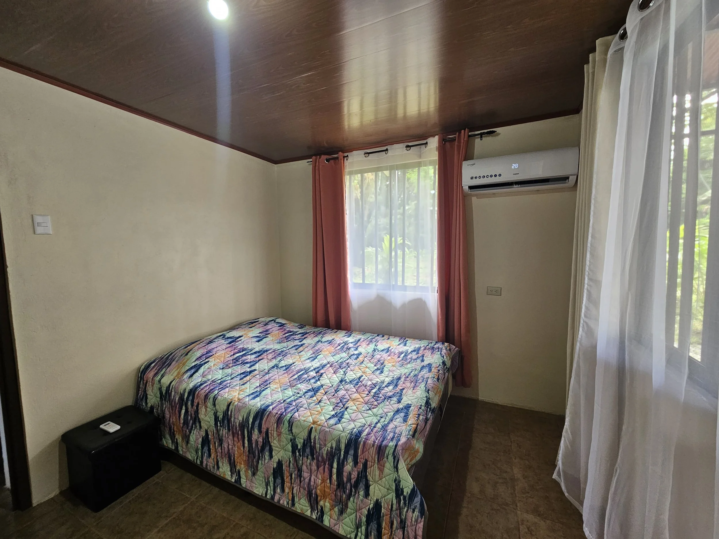 A bedroom with a bed covered in a colorful patterned bedspread, a window with white sheer curtains and pink drapes, an air conditioner on the wall, and a small black nightstand next to the bed.