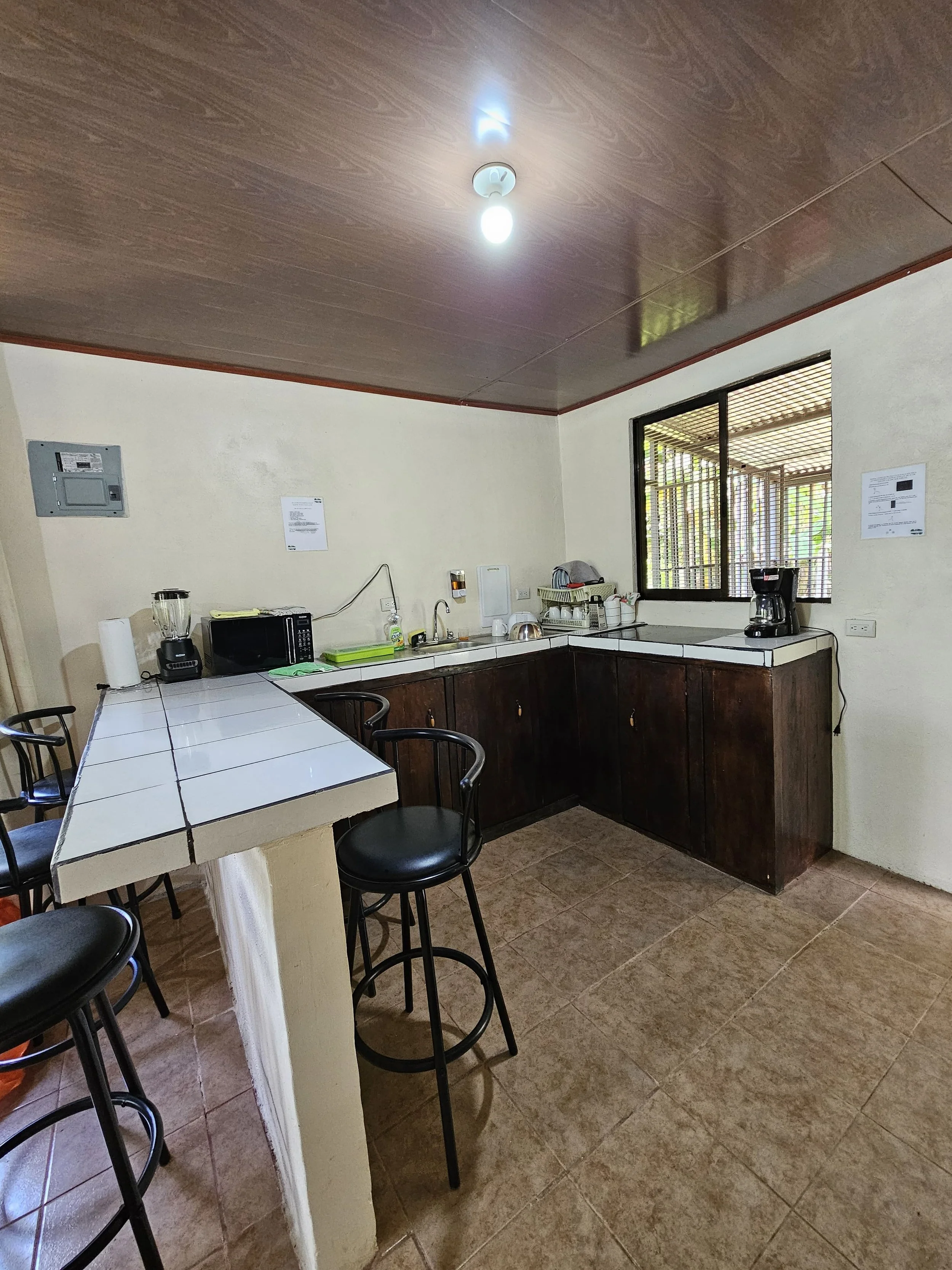 A small kitchen with a tiled breakfast bar, dark wooden cabinets, white countertops, and various appliances including a microwave, blender, coffee maker, and dish drying rack. A window with wooden blinds allows natural light into the room.