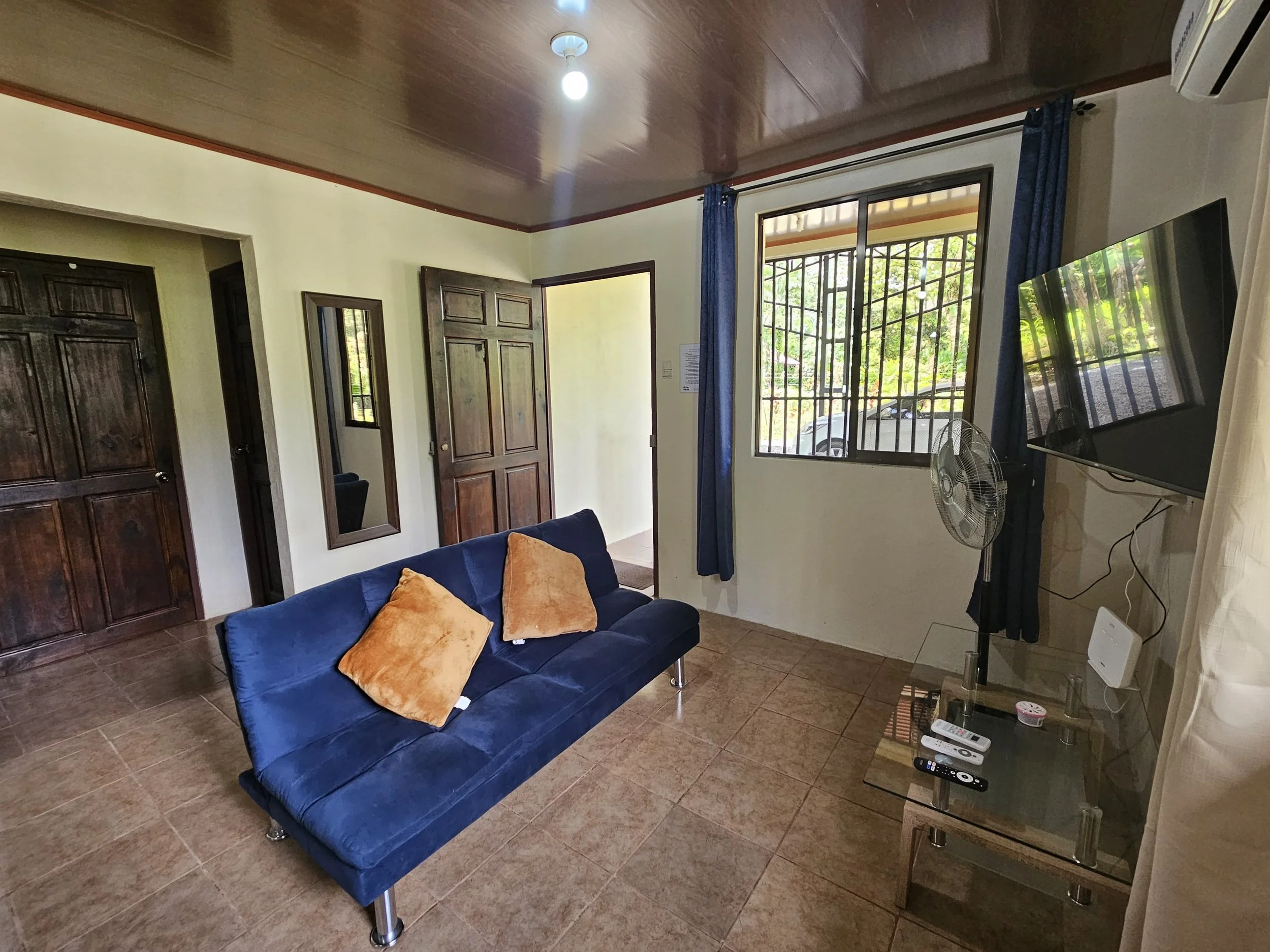 Living room with a blue sofa with two tan pillows, a flat-screen TV on the wall, a glass TV stand with remote controls, a standing fan, and an open window with blue curtains showing outside greenery.