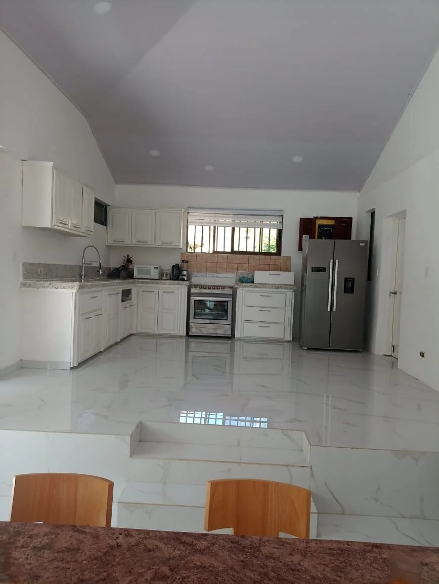 Empty kitchen with white cabinets, stainless steel refrigerator, and marble floor, viewed from a dining area.