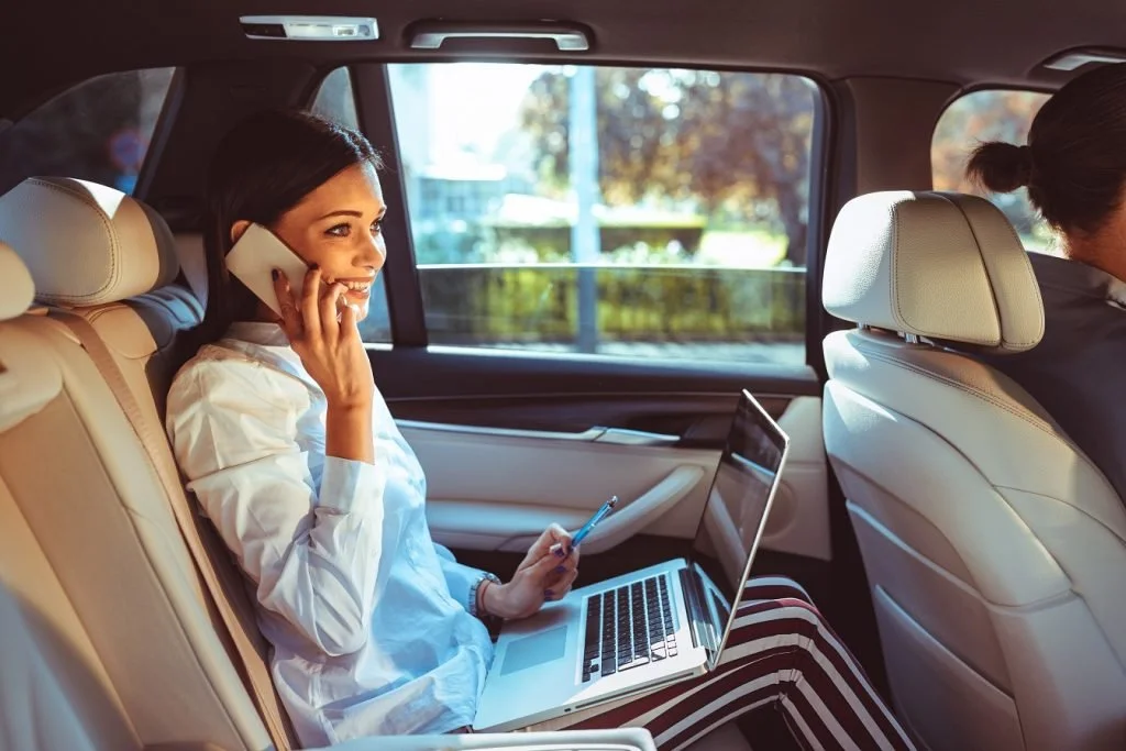 Woman sitting in the backseat of a car, talking on her cellphone, using a laptop and holding a pen.