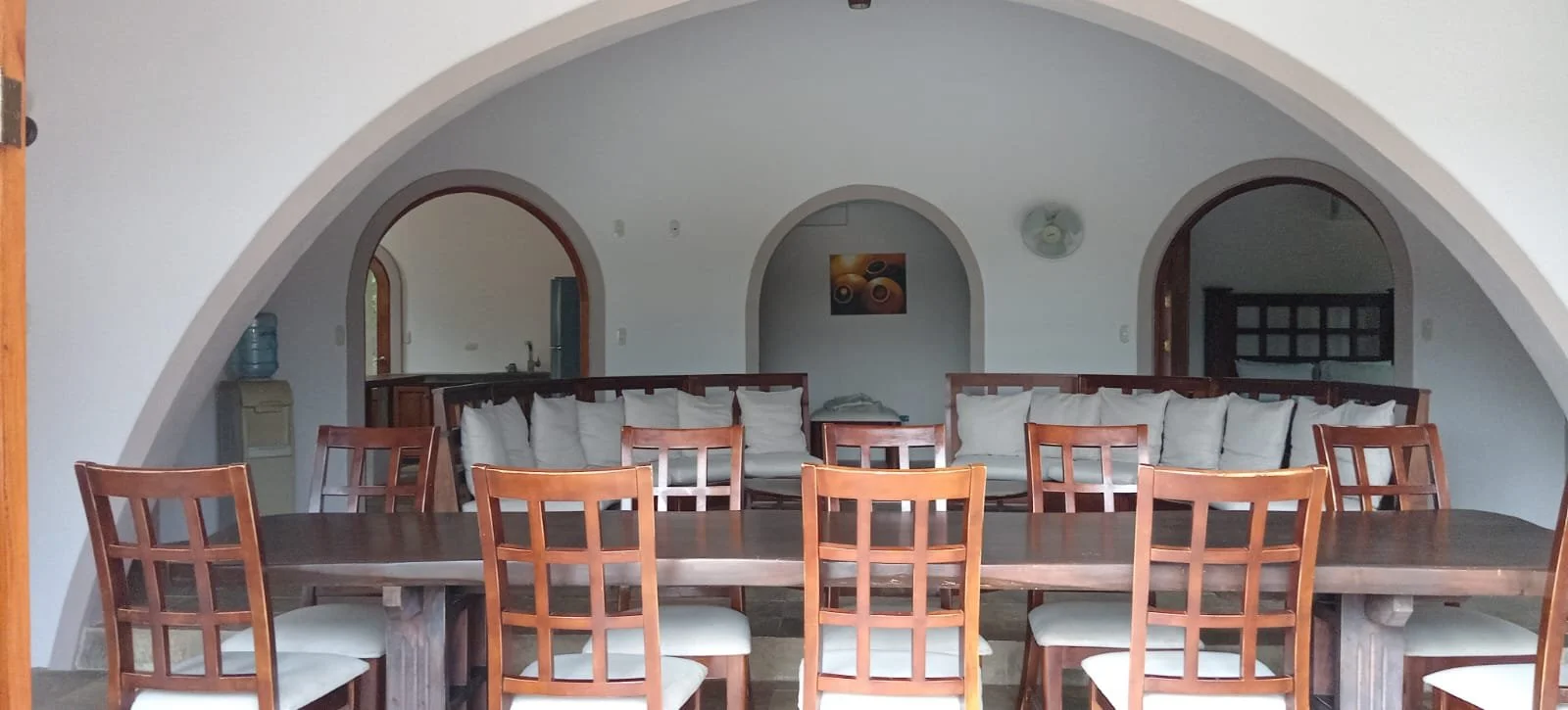 Dining room with a long wooden table, wooden chairs with white cushions, a white corner couch with pillows, and arched doorways leading to other rooms