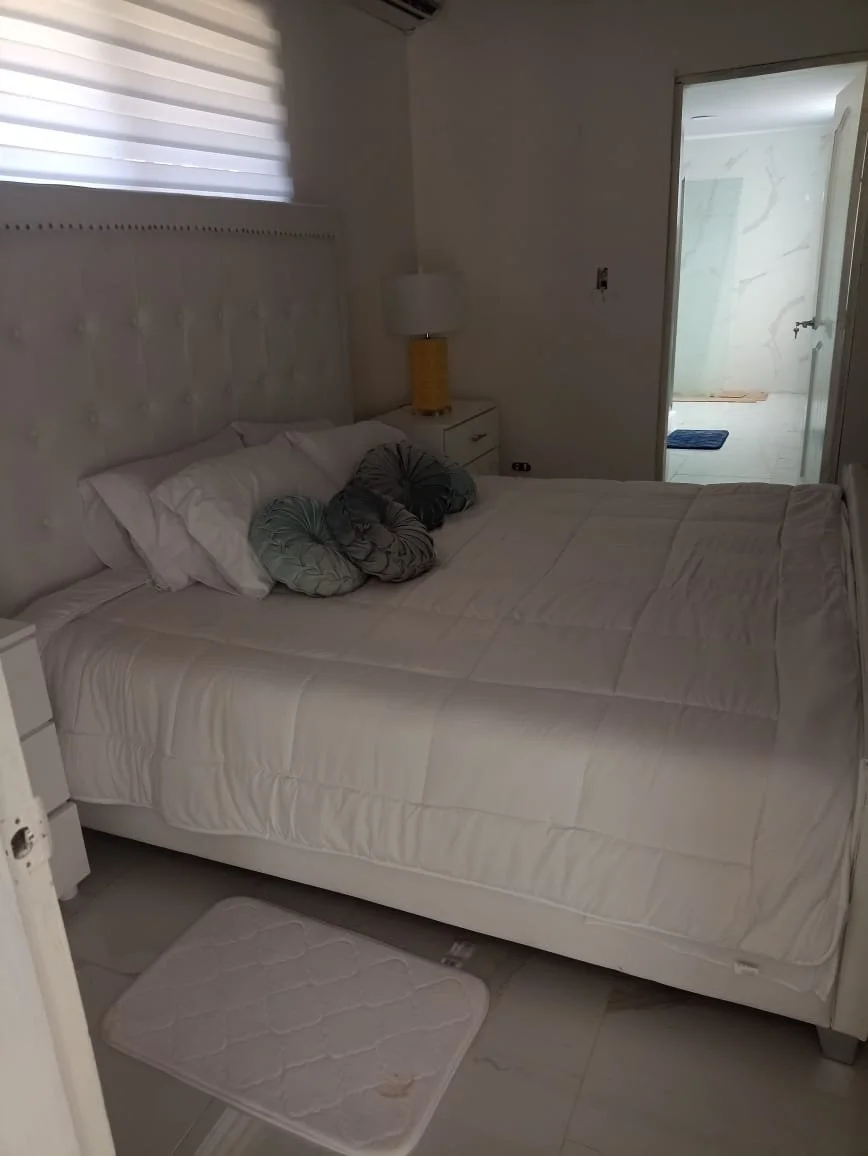 A neatly made bed with white bedding and two gray decorative pillows, a white nightstand with a yellow lamp, and a white bathmat on the floor. There is a window with white blinds and an open door leading to a tiled bathroom.