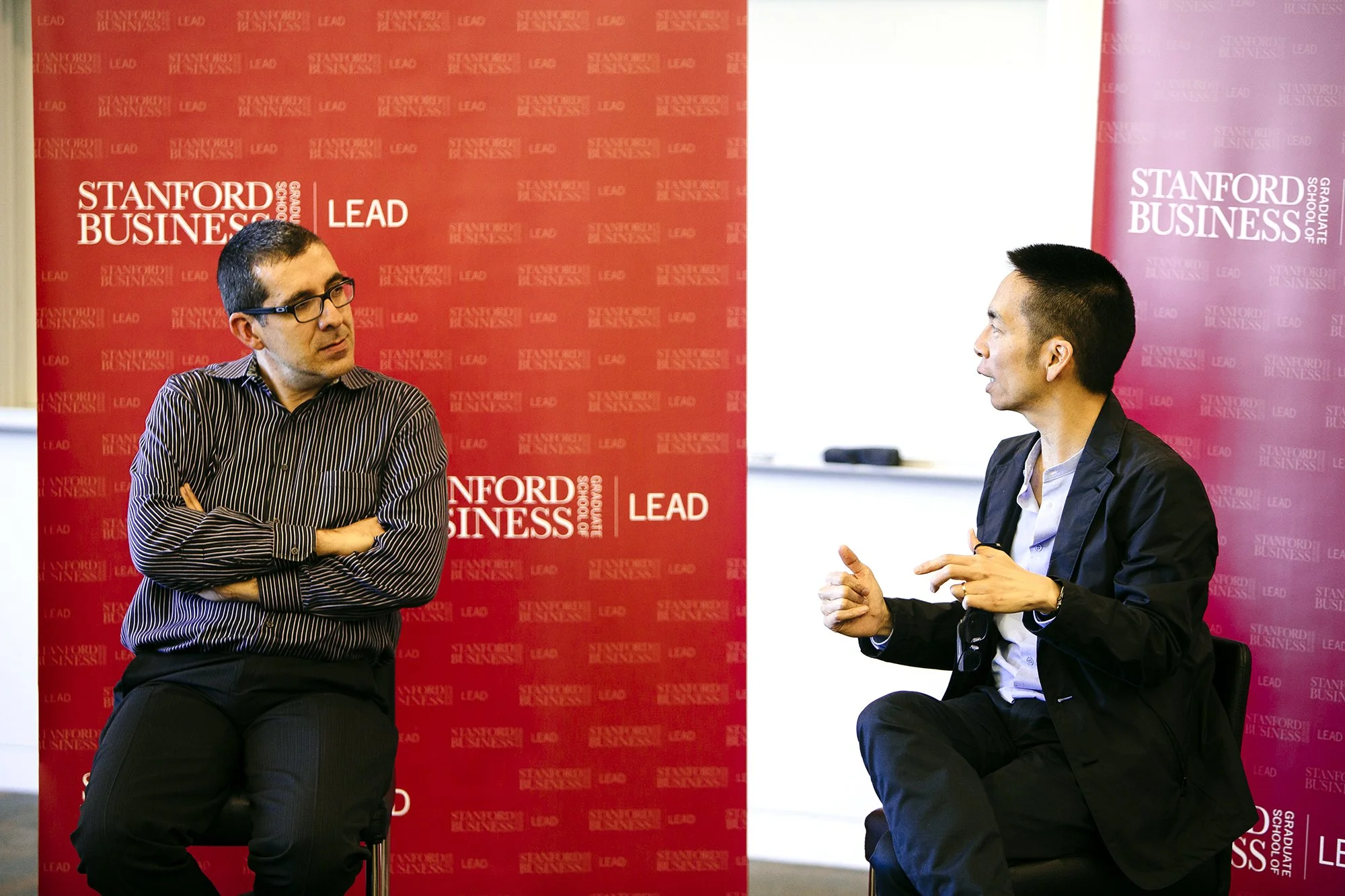 Two graduate students engaged in conversation during a discussion or interview at Stanford Graduate School of Business, with branded cardinal red Stanford LEAD logo.