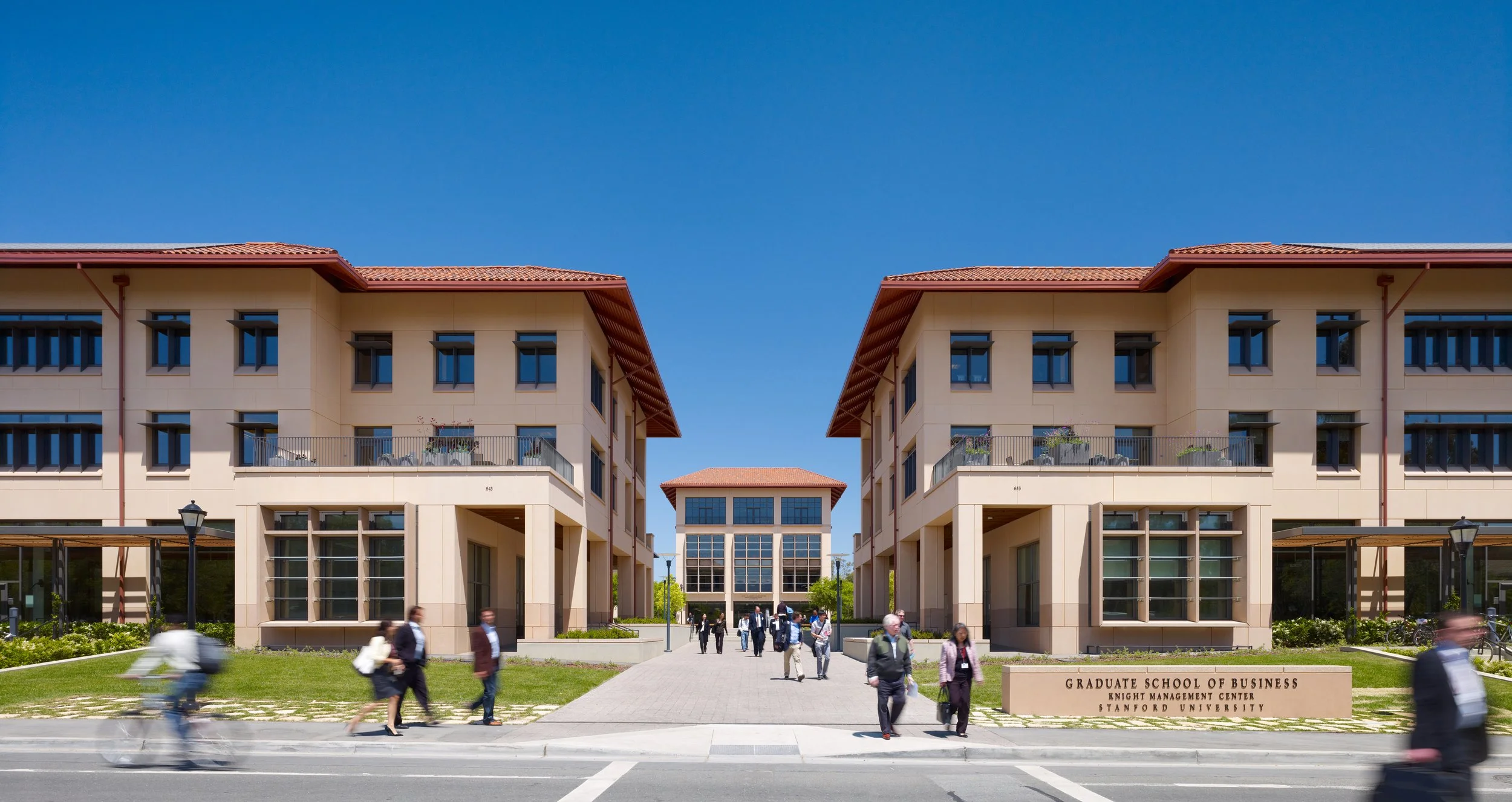 View of Stanford University's Graduate School of Business campus with modern buildings, people walking, and a clear blue sky using low shutter photography.
