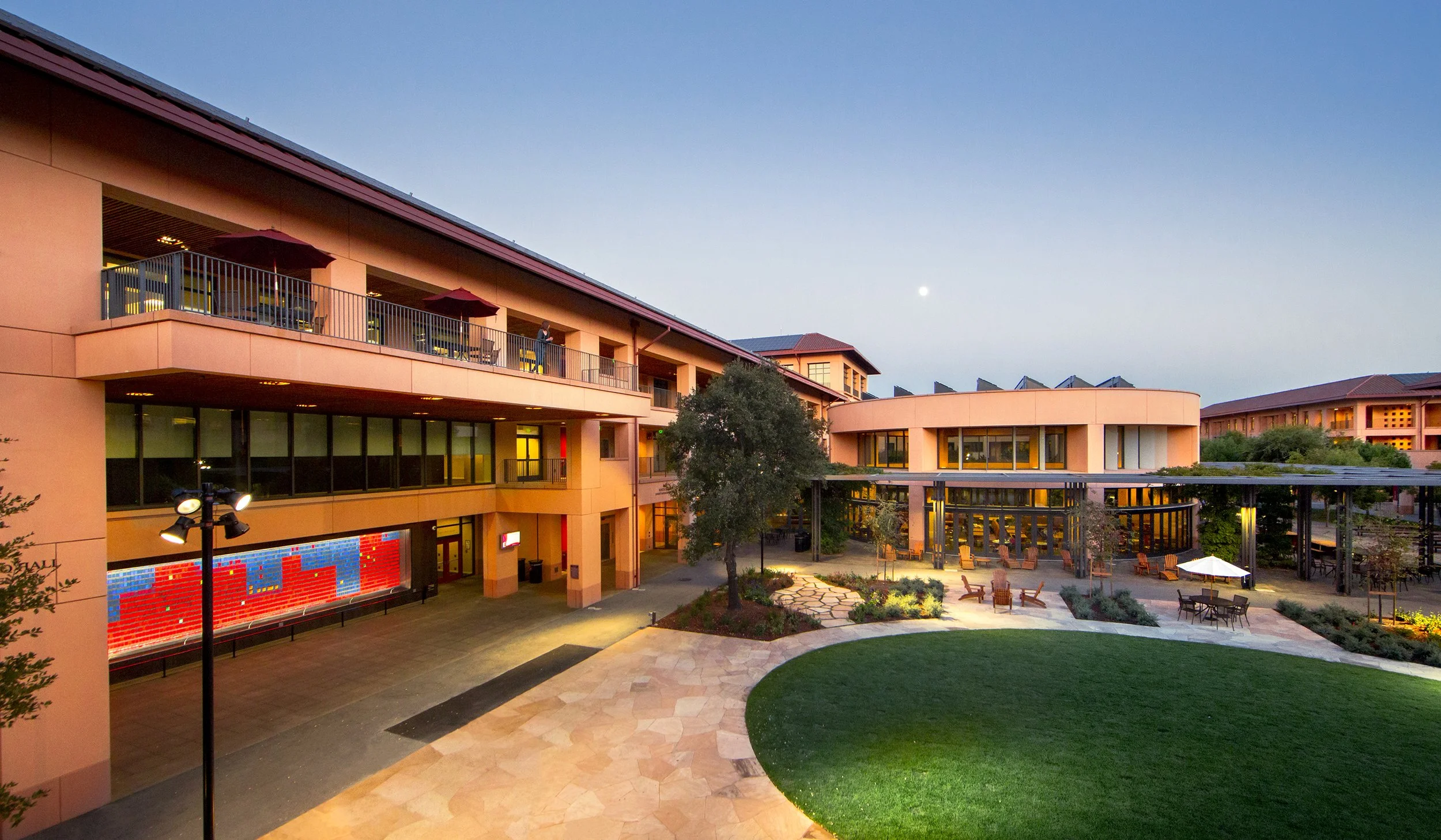 Exterior Stanford Graduate School of Business campus at dusk of a modern multi-story building with a courtyard, outdoor seating, and landscaped gardens.