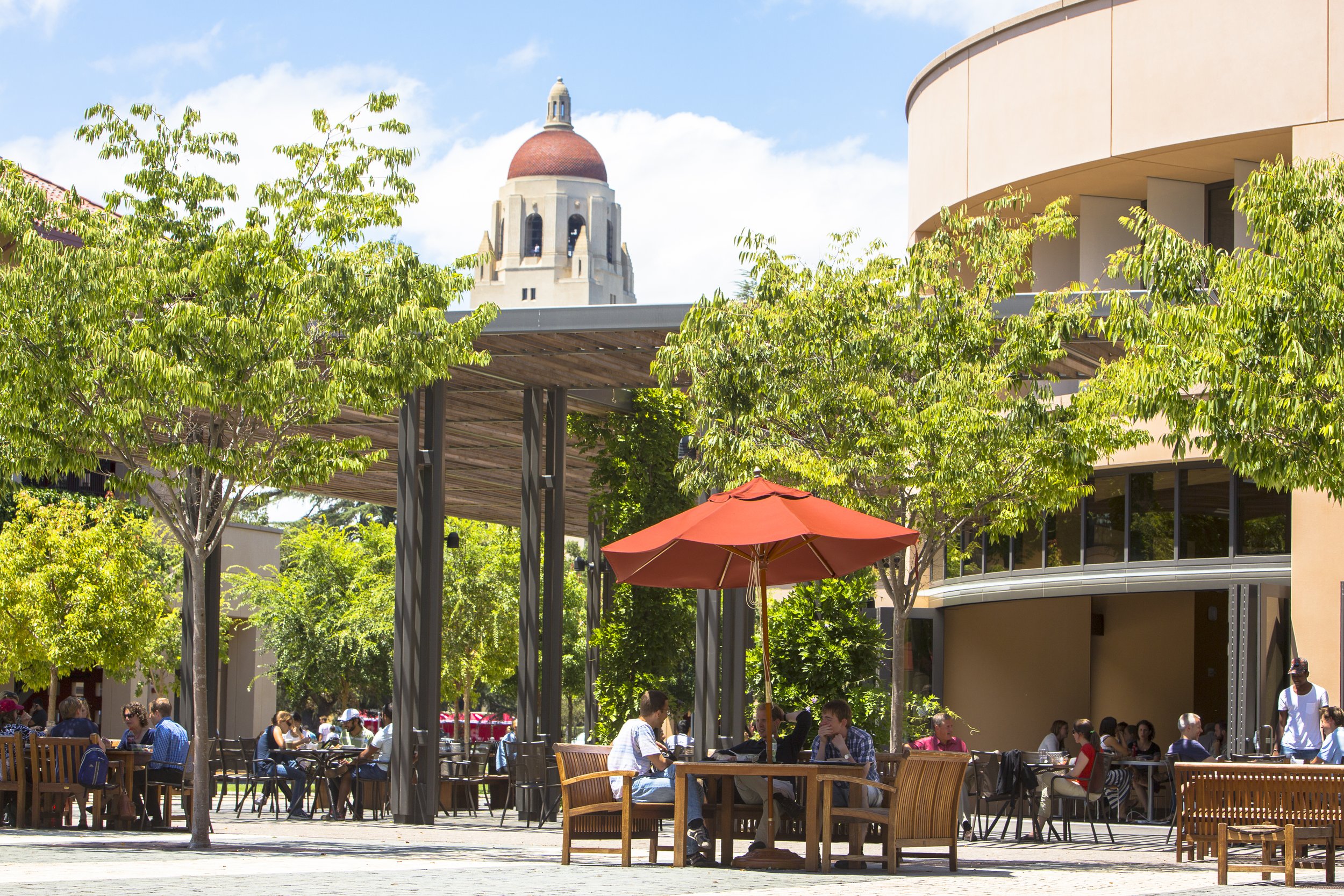 People dining outdoors at Arbuckle Dining Pavilion on Knight Management Center Stanford GSB campus on tables with umbrellas with trees and Hoover Tower in the background.