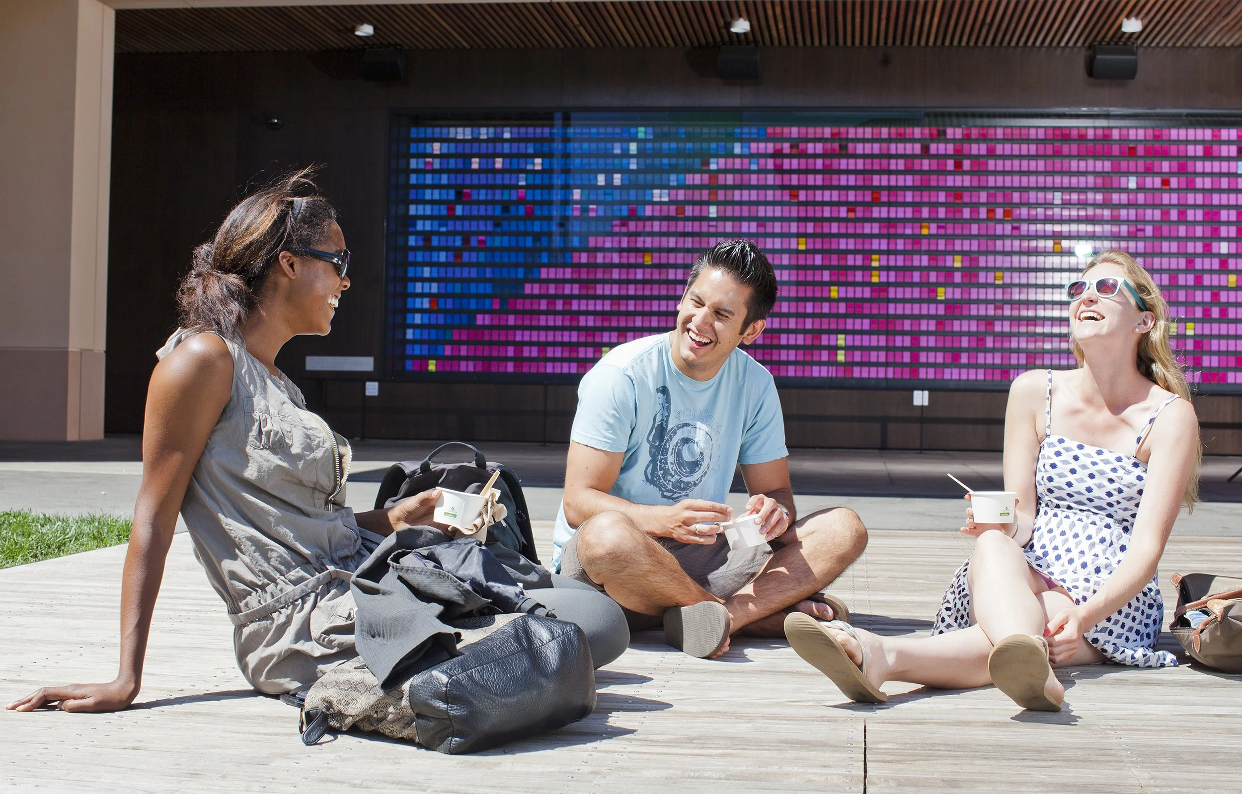 Three Stanford GSB students sitting outside in front of "Monument to Change as It Changes" kinetic art installation, smiling and talking during town hall meeting on campus.