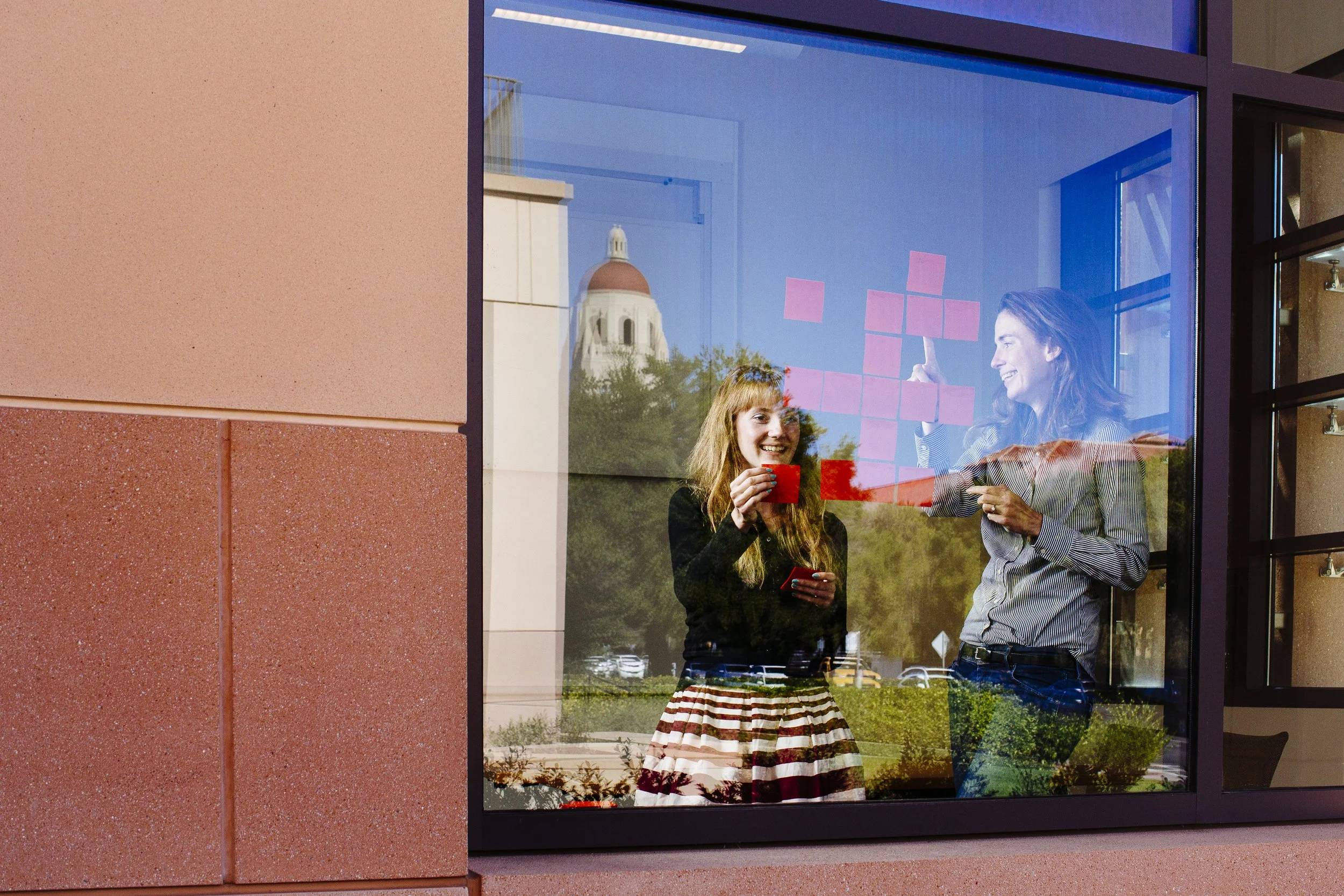 Natalie Rumao and Toni Bird are seen through a glass window, smiling and engaging in a brainstorm using sticky notes on the windows of the Stanford GSB campus building with a reflection of buildings, Hoover Tower, and trees.