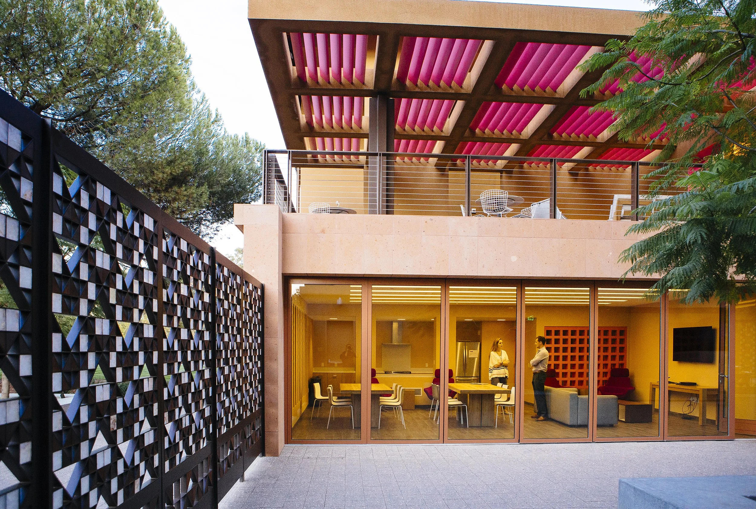 Stanford GSB brand refresh inspiration architecture image. Modern building with large glass windows, an interior space with brightly colored chairs and tables, and people inside, surrounded by trees and greenery at Highland Hall.