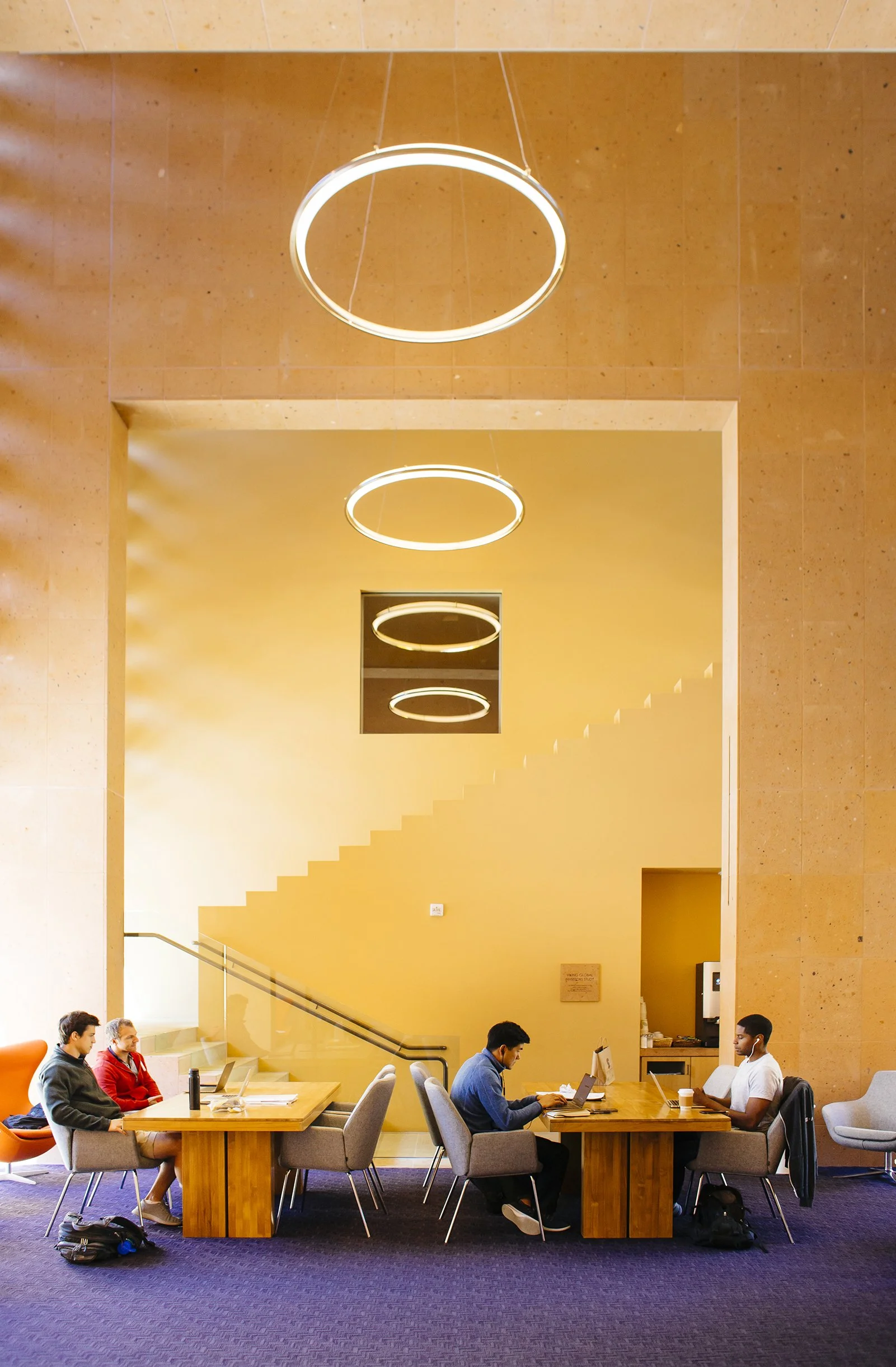 Stanford GSB brand refresh inspiration campus architecture image. Modern indoor space with a large wooden table and chairs, people working on laptops, and circular ceiling lights on a yellow-orange wall.