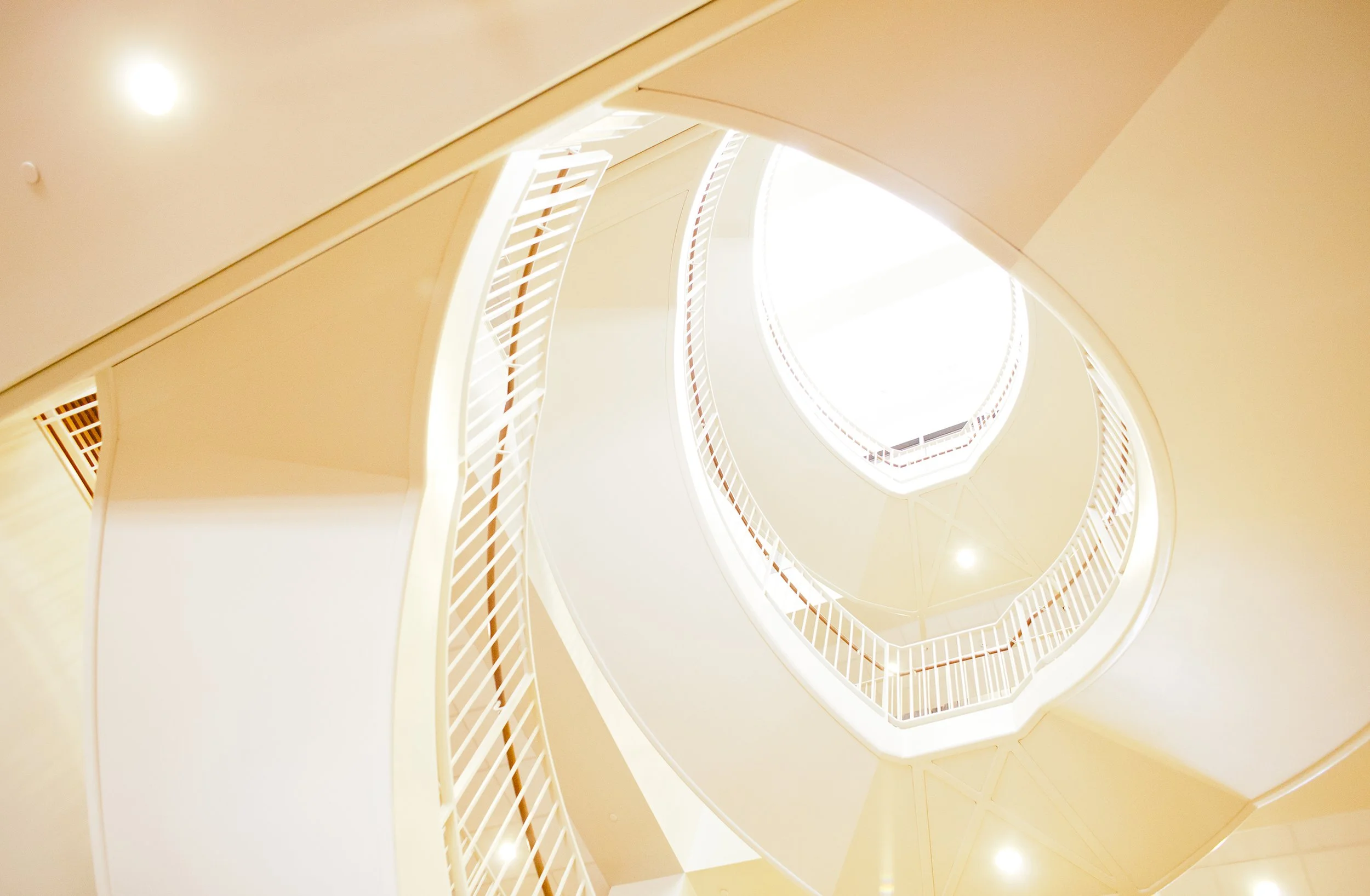 Stanford GSB brand refresh inspiration architecture image. Looking up at a spiral staircase from the bottom of library, with bright light at the top and white railings surrounding the open space.