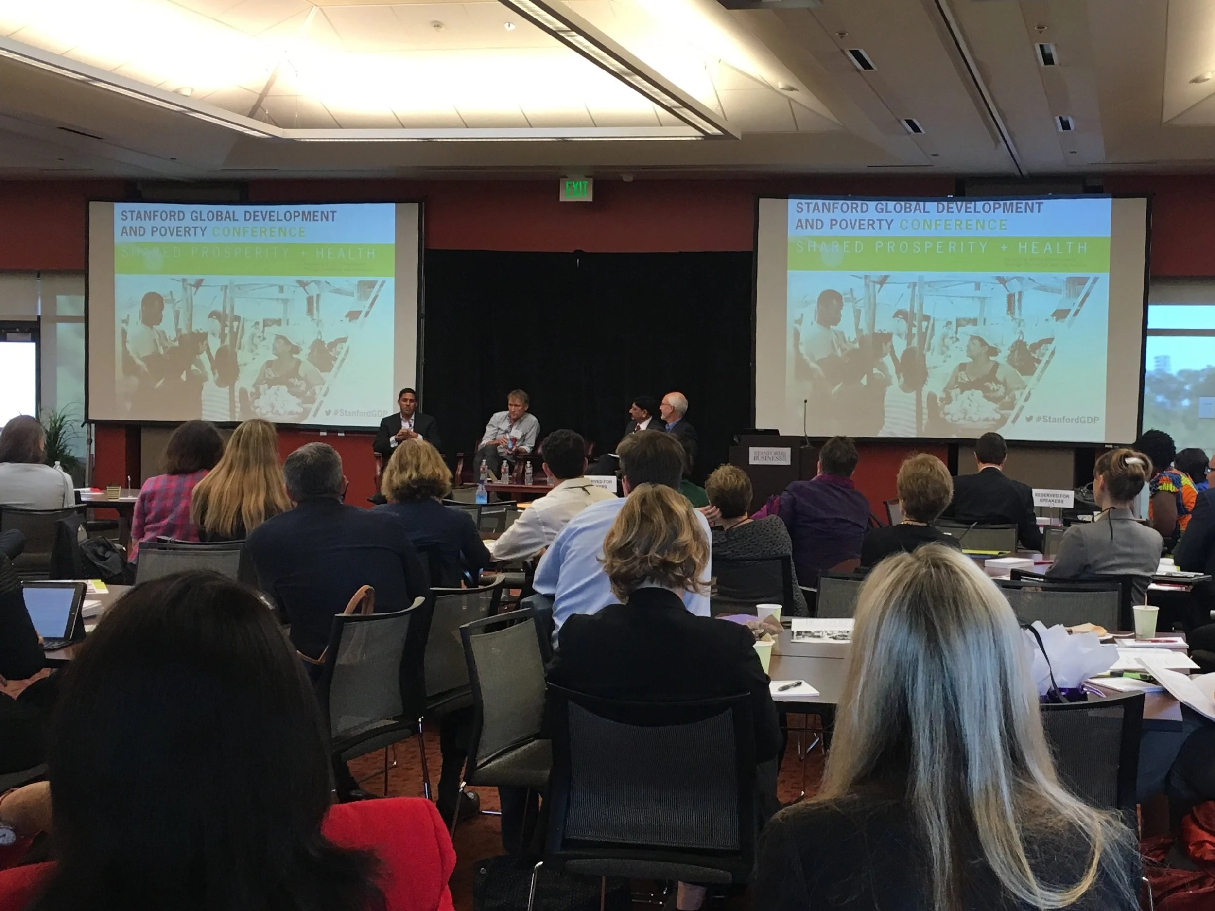 A conference room filled with seated attendees facing a stage with two large screens displaying the title 'Stanford Global Development and Poverty Conference.' Three panelists are seated on the stage.