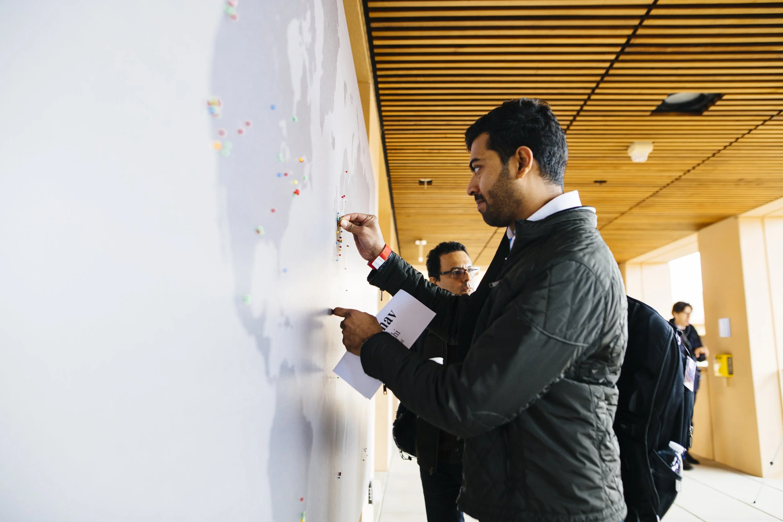 A man with short black hair, wearing a black jacket, is pinning a colorful pushpin into a Stanford Graduate School of Business LEAD program, large printed interactive map.