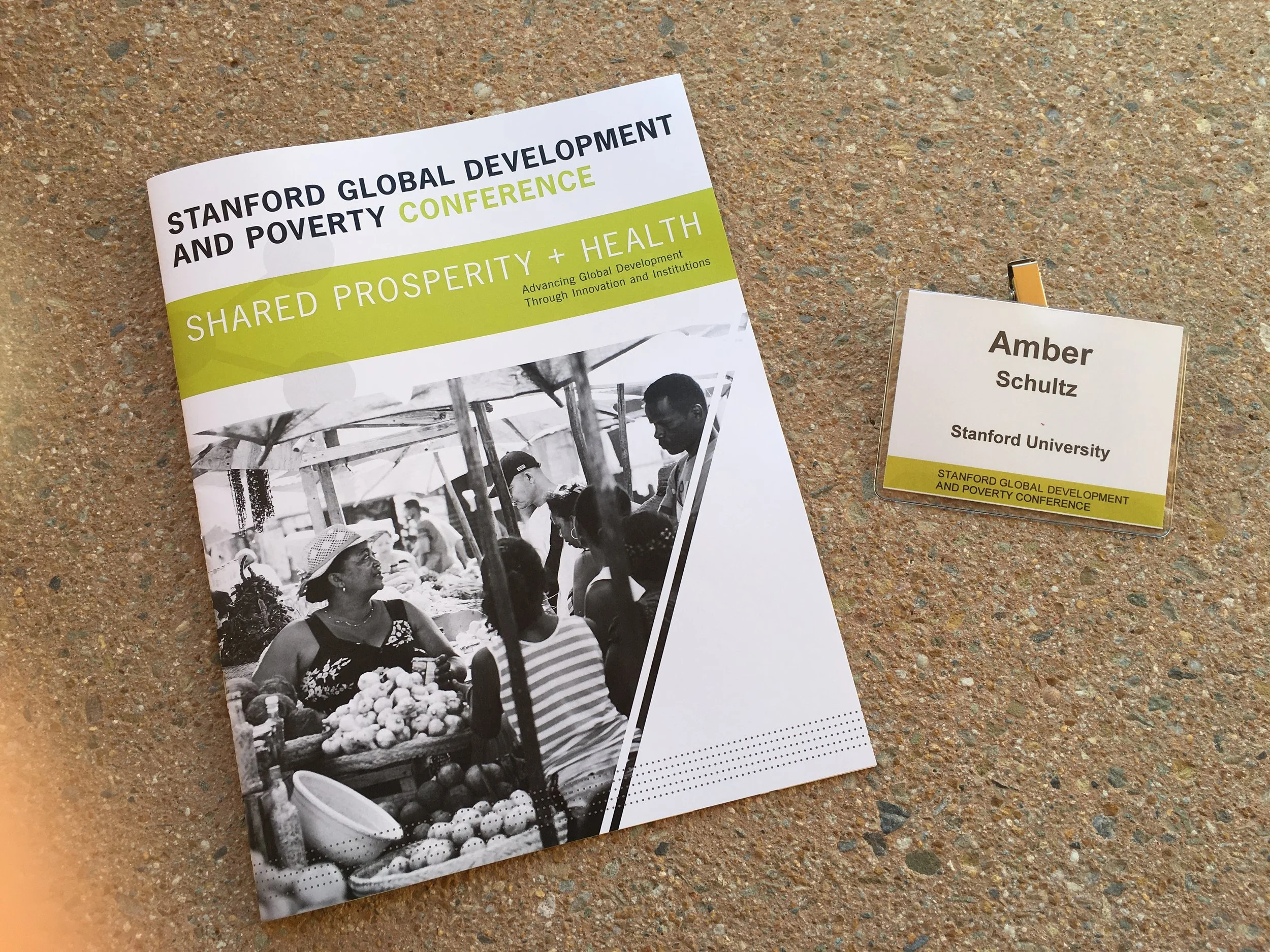 Conference brochure and name tag on a textured surface. The brochure reads 'Stanford Global Development and Poverty Conference, Shared Prosperity + Health, Advancing Global Development Through Innovation and Institutions'.