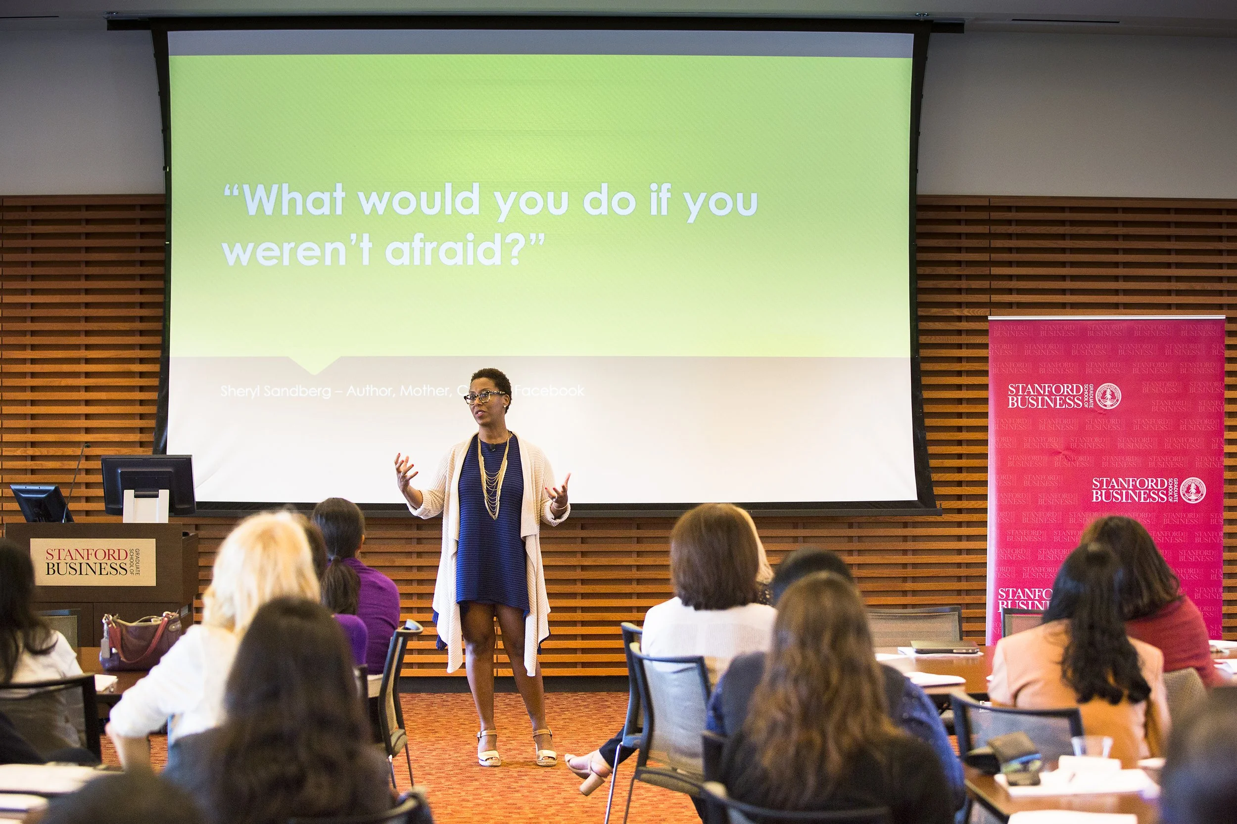 A woman is speaking at a conference at Stanford Graduate School of Business, with GSB branded banners and a large presentation slide displaying the quote 'What would you do if you weren't afraid?' The audience is seated, listening to the speaker.
