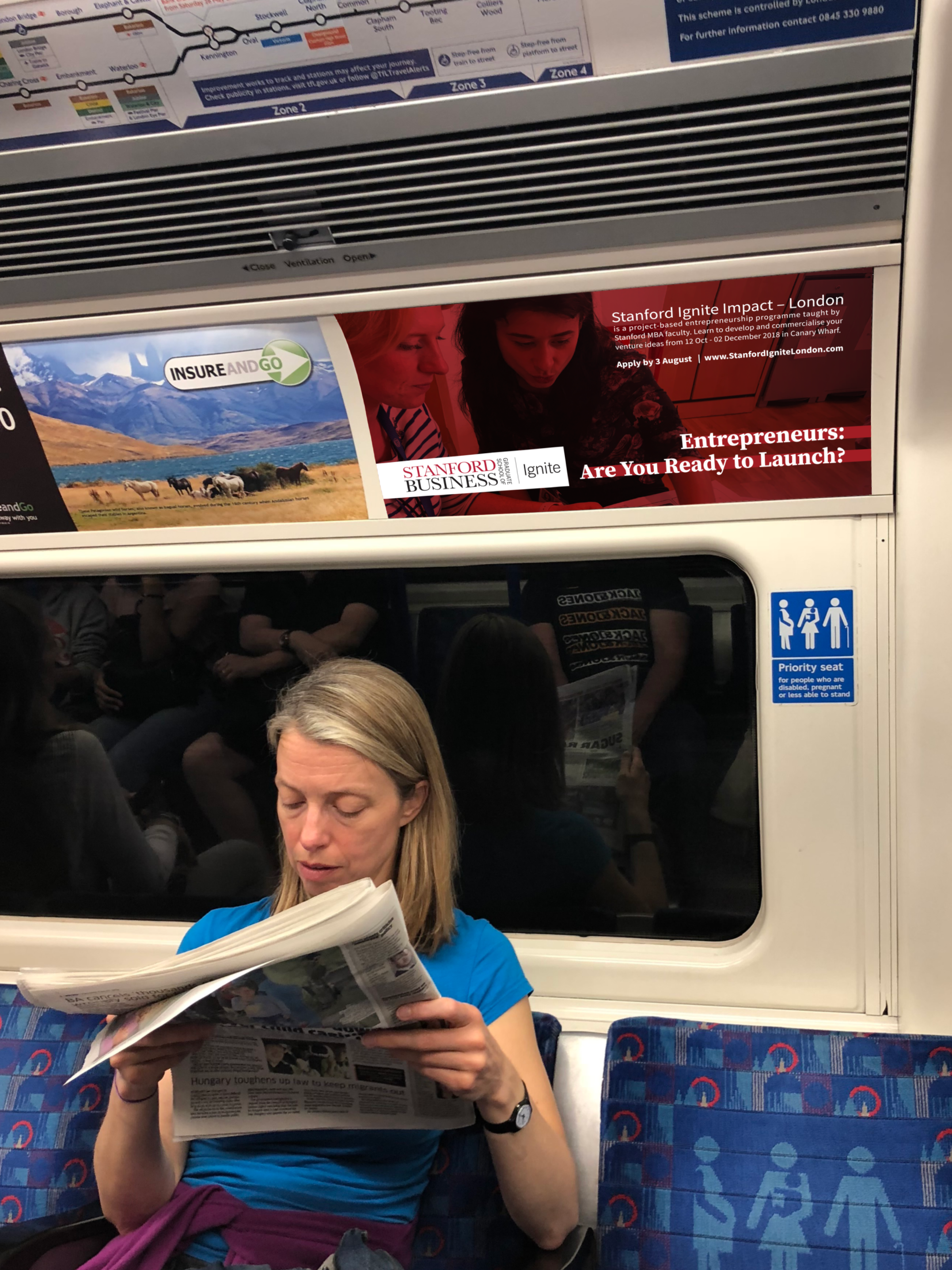 A passenger of the London Tube, reading a newspaper. Above her, an advertisement promoting Stanford Ignite: Impact in London.