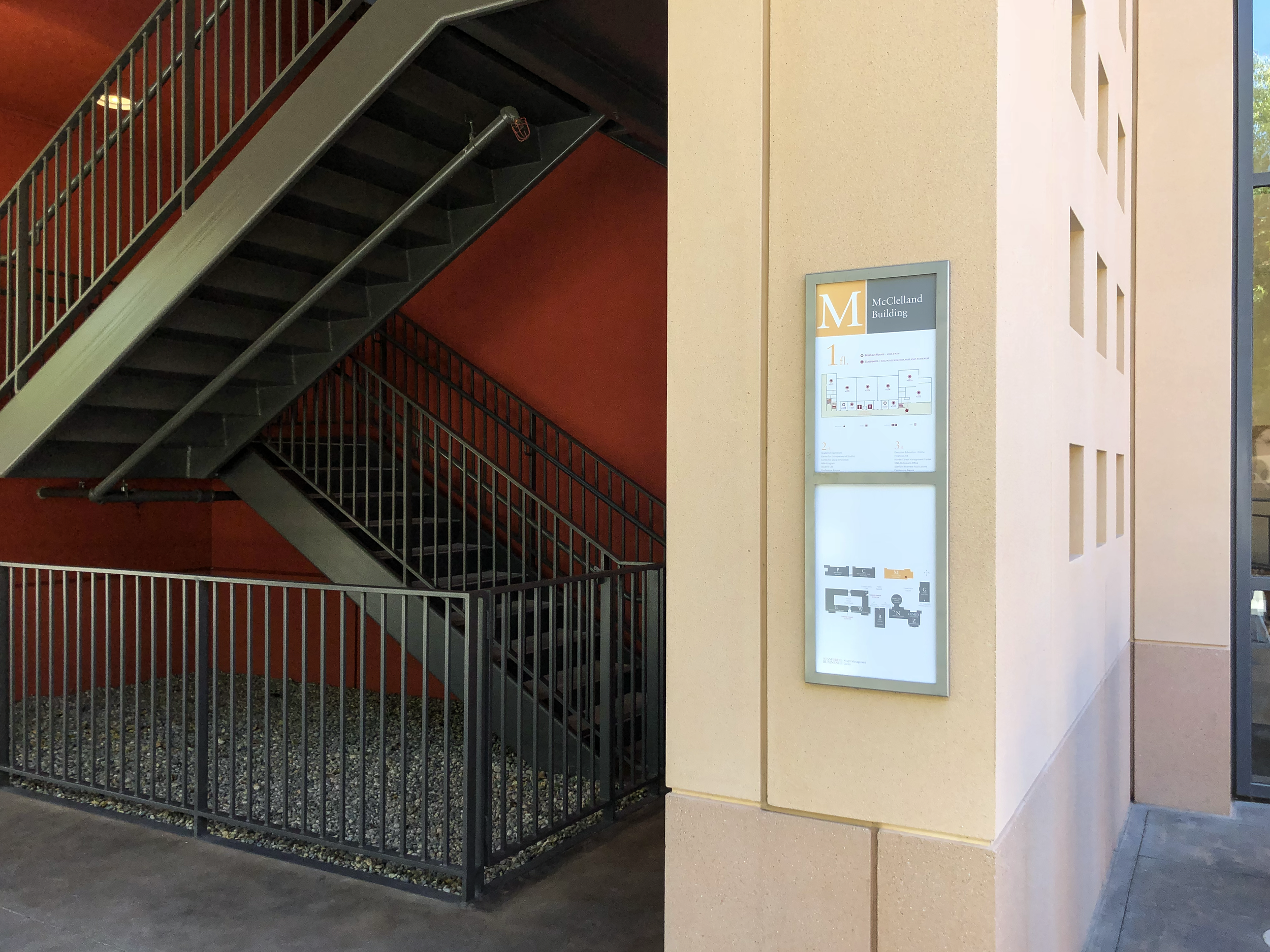 Wayfinding at Stanford Graduate School of Business showing a metal staircase, a yellow wall with small square cutouts, and a directory sign with a branded building map.