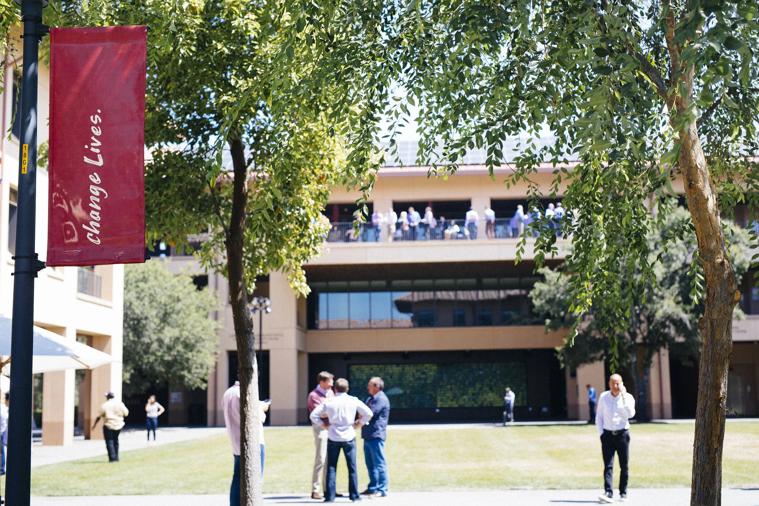 A red vertical banner with white text that reads 'Change Lives' is hanging from a lamp post at Stanford GSB. There are multiple trees and a crowd of people walking and standing around campus.