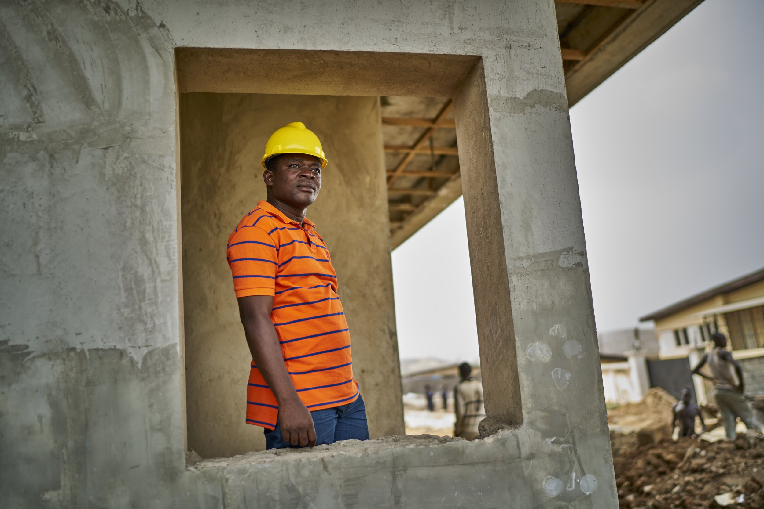 Award Winning Stanford Seed image: A man in an orange and blue striped shirt and a yellow safety helmet stands in front of a concrete building under construction, looking into the distance in Africa.