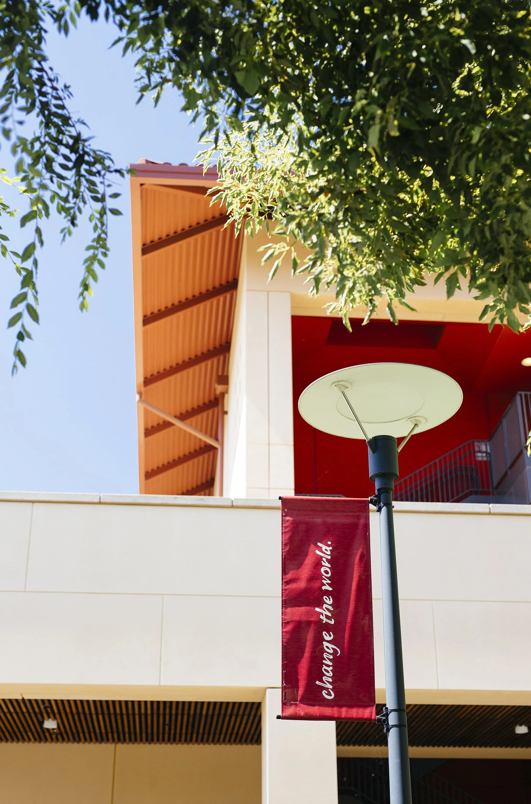 A red vertical banner with white text that reads 'Change the World' is hanging from a lamp post at Stanford GSB. Green tree branches and leaves are visible in the upper part of the image, and the sky is clear and blue.