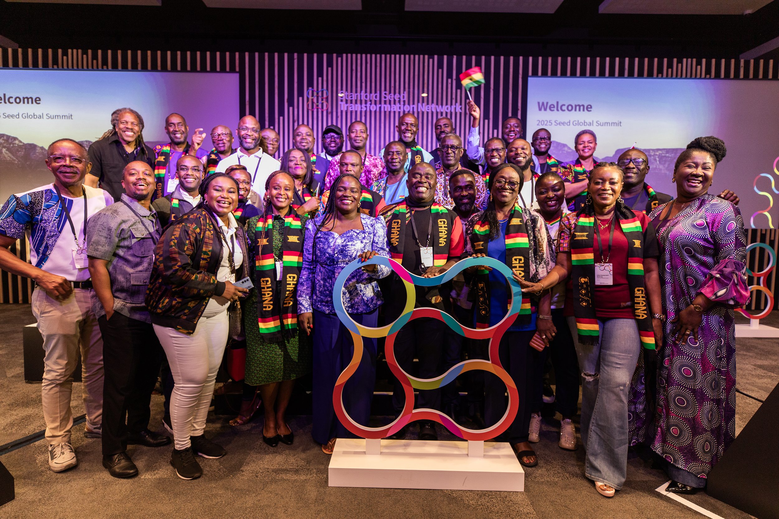 Group of diverse people smiling at a Stanford Seed Network event on stage, some wearing scarves with Ghana's flag colors, standing behind a colorful sculpture and a large screen displaying 'Welcome 2025 Seed Global Summit'.