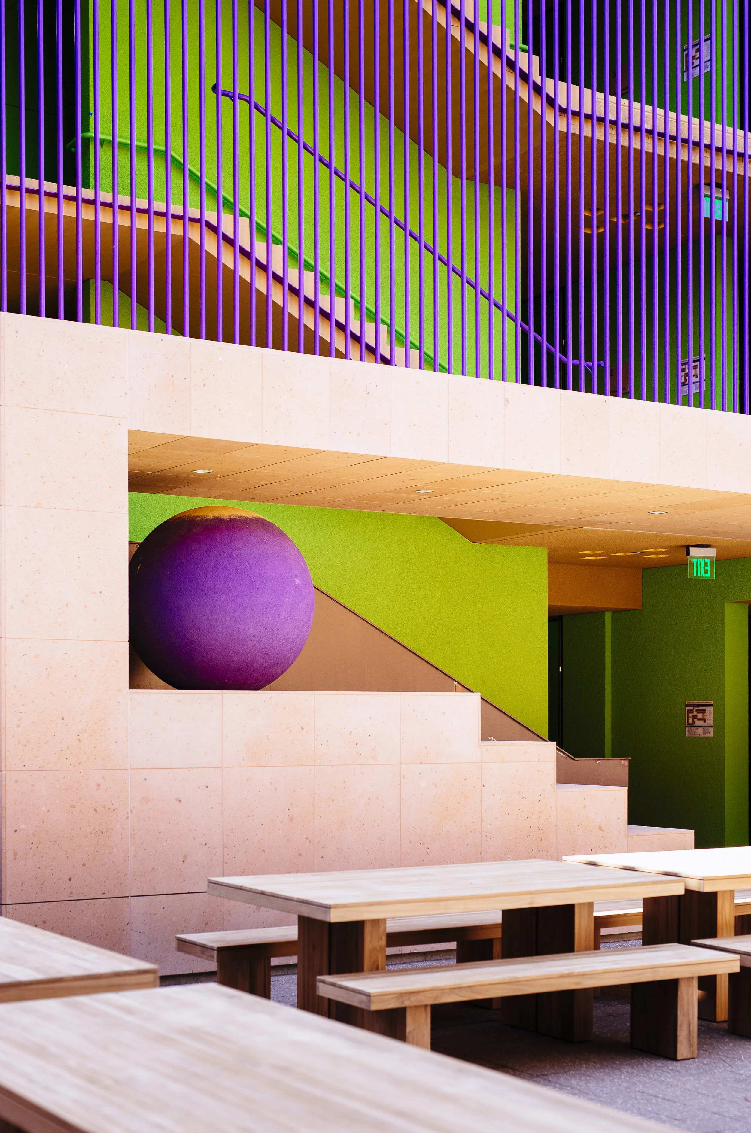 Stanford GSB brand refresh inspiration architecture image. Interior of a modern space with a purple large sphere, wooden tables and benches, a staircase with purple vertical bars, green walls, and exit signs of Highland Hall.