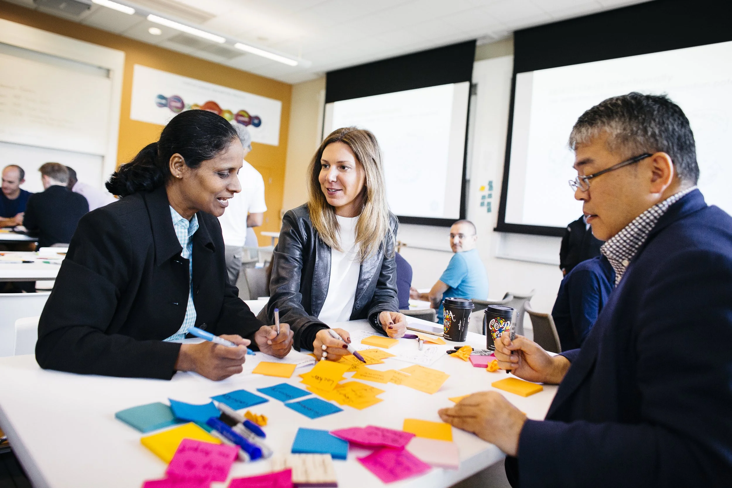 Group of diverse professionals at Stanford Graduate School of Business engaged in a collaborative work session with sticky notes and pens, set in a modern office space with presentation screens.