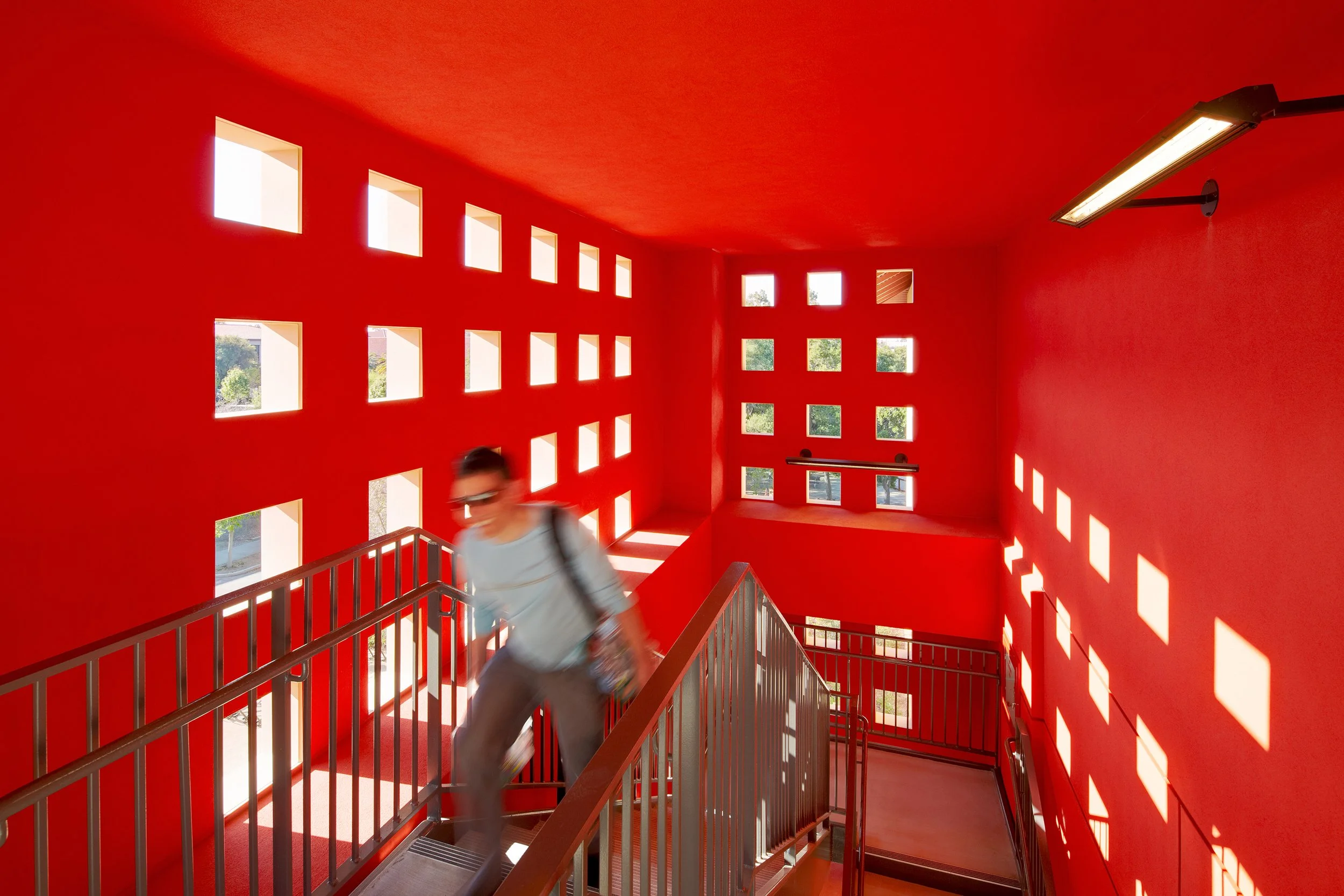 Award Winning Stanford GSB image: A bright red-painted interior stairwell with square windows allowing sunlight to cast shadows on the walls. A person with glasses and a backpack is ascending the stairs in low shutter photography.