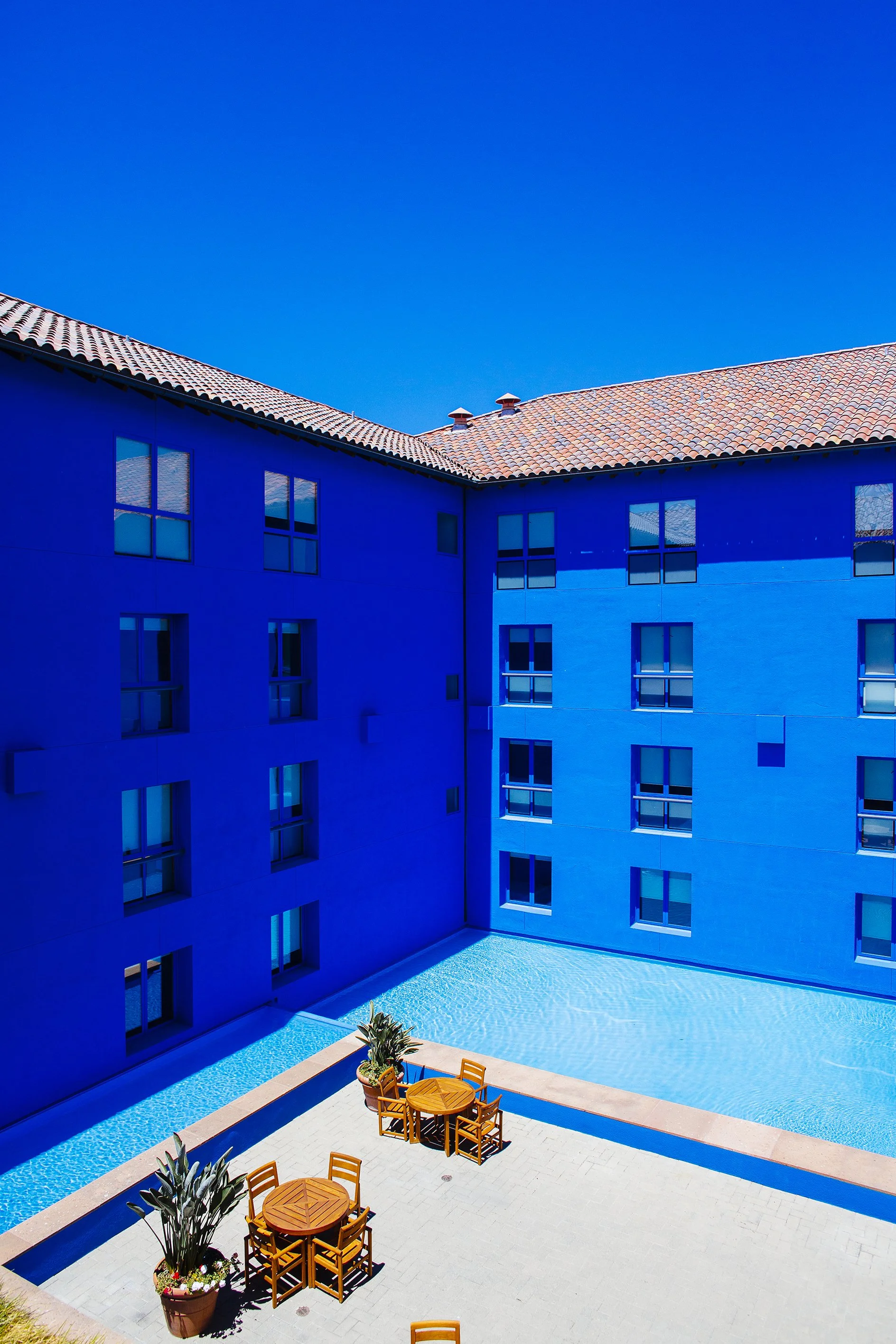 Stanford GSB brand refresh inspiration architecture image. The image shows a courtyard of a blue campus building with a swimming pool. Olive trees with potted plants and wooden chairs are arranged around the pool area, under a clear blue sky.