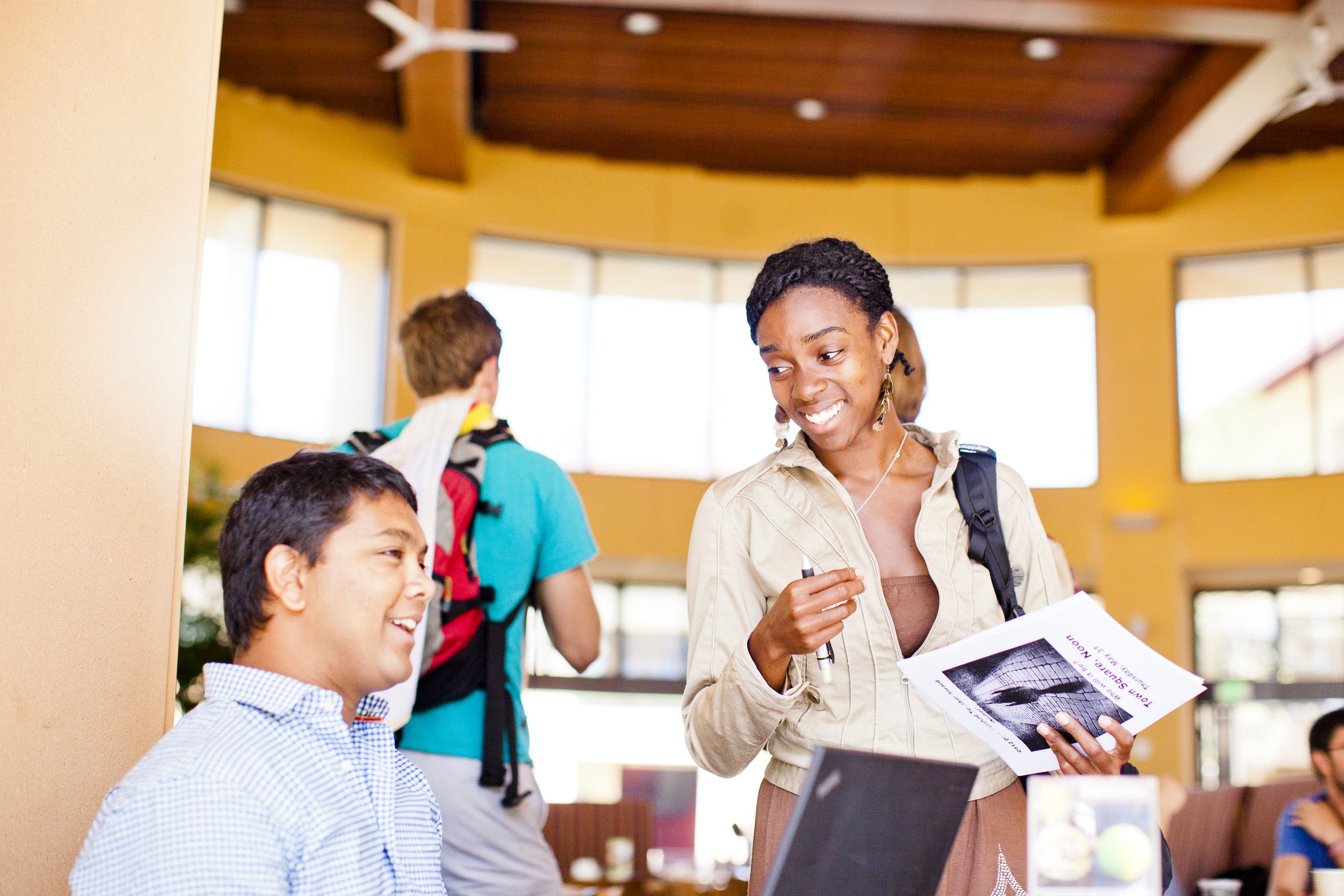A smiling Stanford Graduate School of Business student holding papers and a pen, engaging in conversation with a young man sitting with a laptop, in Arbuckle Dining Pavilion on Knight Management Center campus.