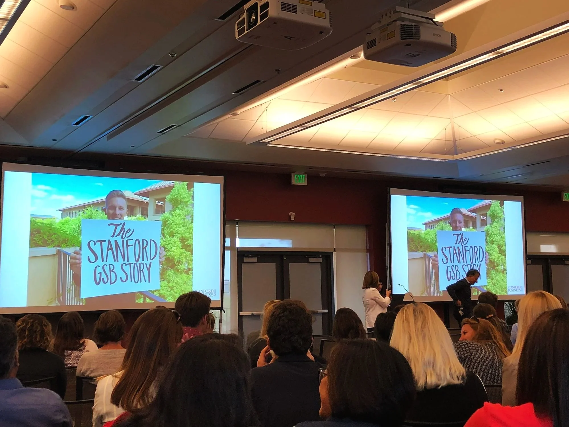 Town hall meeting in conference room filled with staff, two screens display a presentation for 'The Stanford GSB Story,' to rollout the Stanford GSB Brand Refresh
