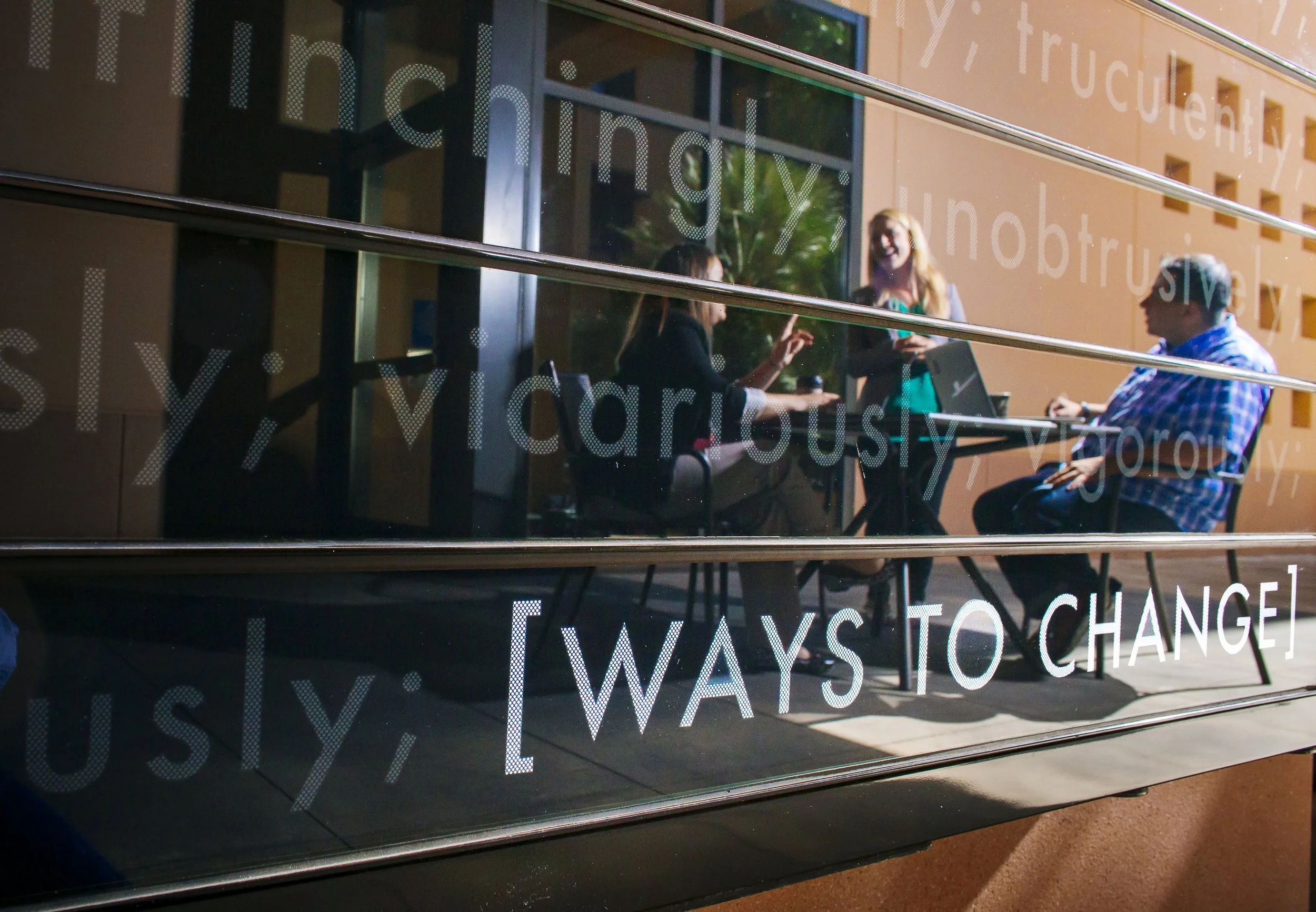 Reflection of people sitting and talking on Change Wall at Stanford Graduate School of Business, visible through reflective glass with white text overlay that reads "[Ways to Change]".