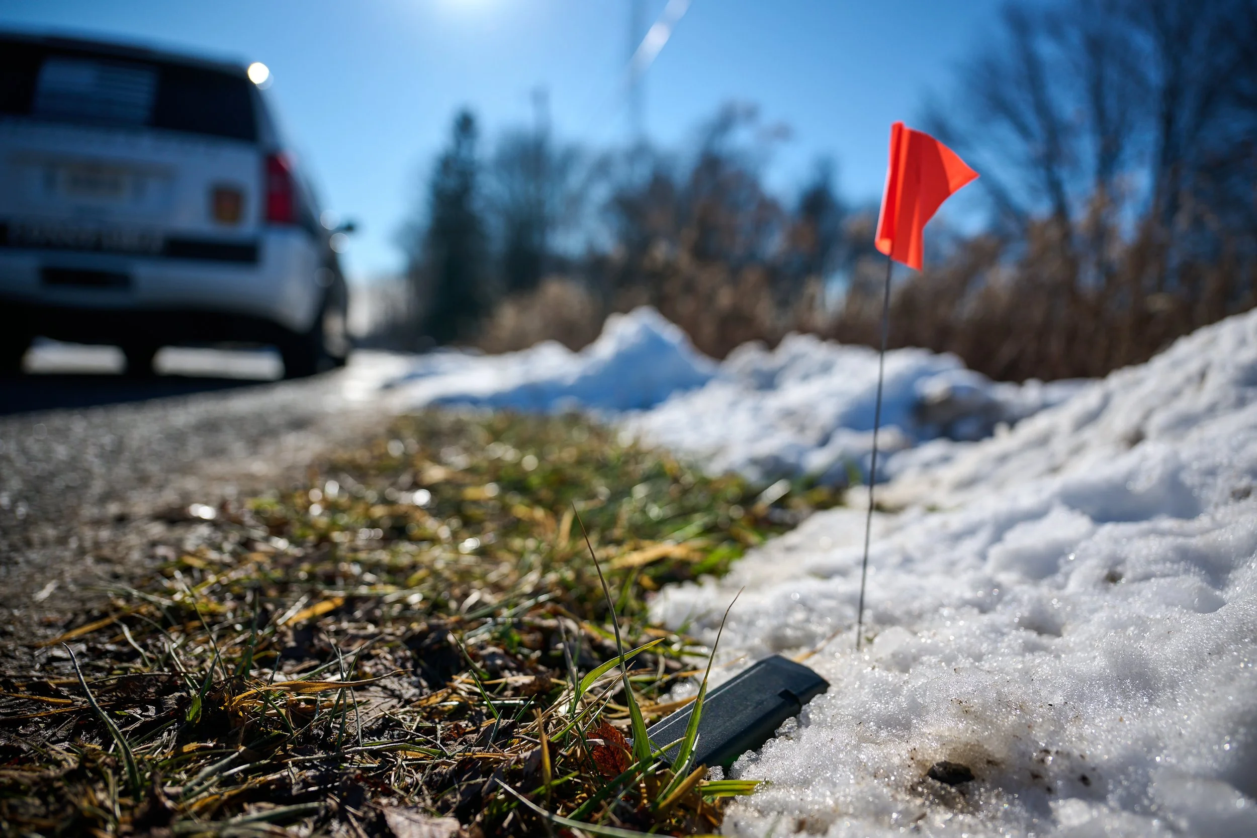 A roadside snow-covered patch with a small red flag on a thin wire, placed to mark a pistol magazine found by K-9 blade