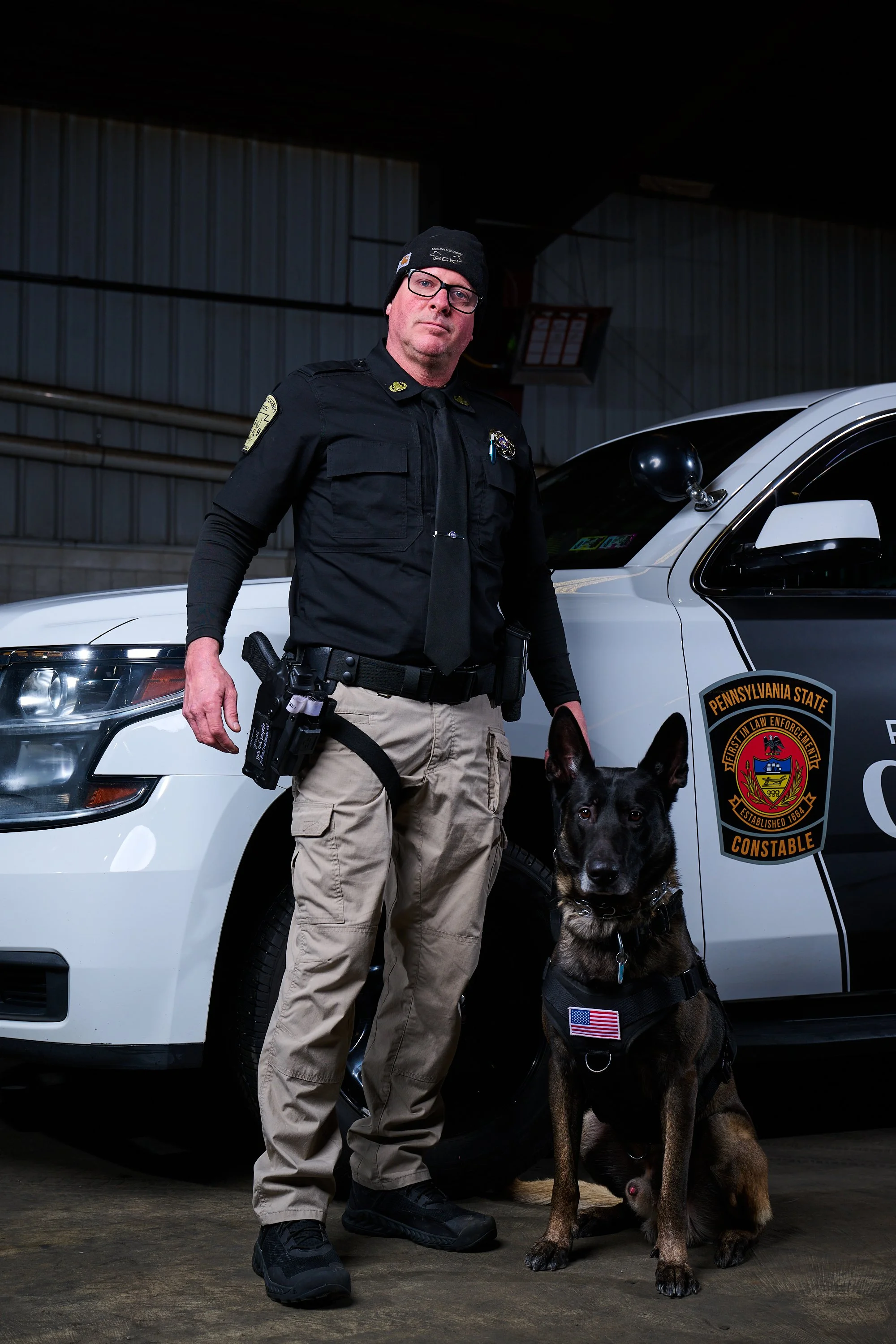 A police officer wearing glasses, a black uniform, and a beanie, standing next to a police car with a K-9 dog, in an indoor garage.