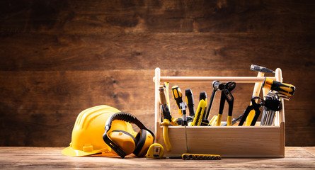 Construction tools and safety equipment including a yellow hard hat, ear protection, and a box of tools on a wooden surface in front of a wood-paneled wall.