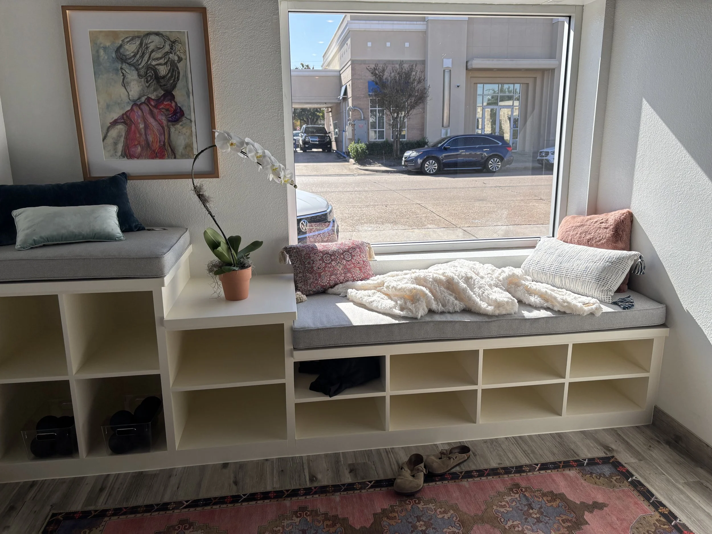 A cozy window seat with cushions and blankets, a potted orchid, and a painting on the wall inside a modern building with large windows and a street view outside.