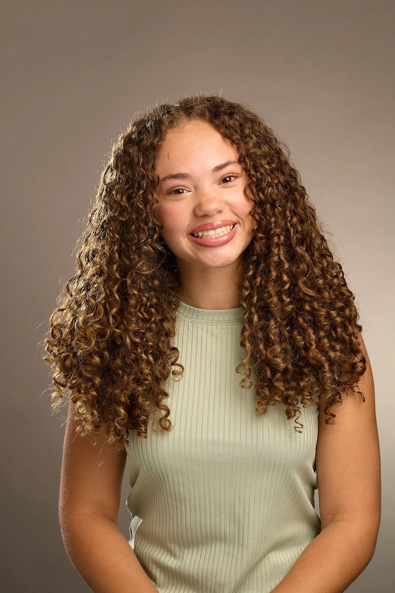 A young girl with curly brown hair wearing a sleeveless, light-colored top, smiling and sticking out her tongue.