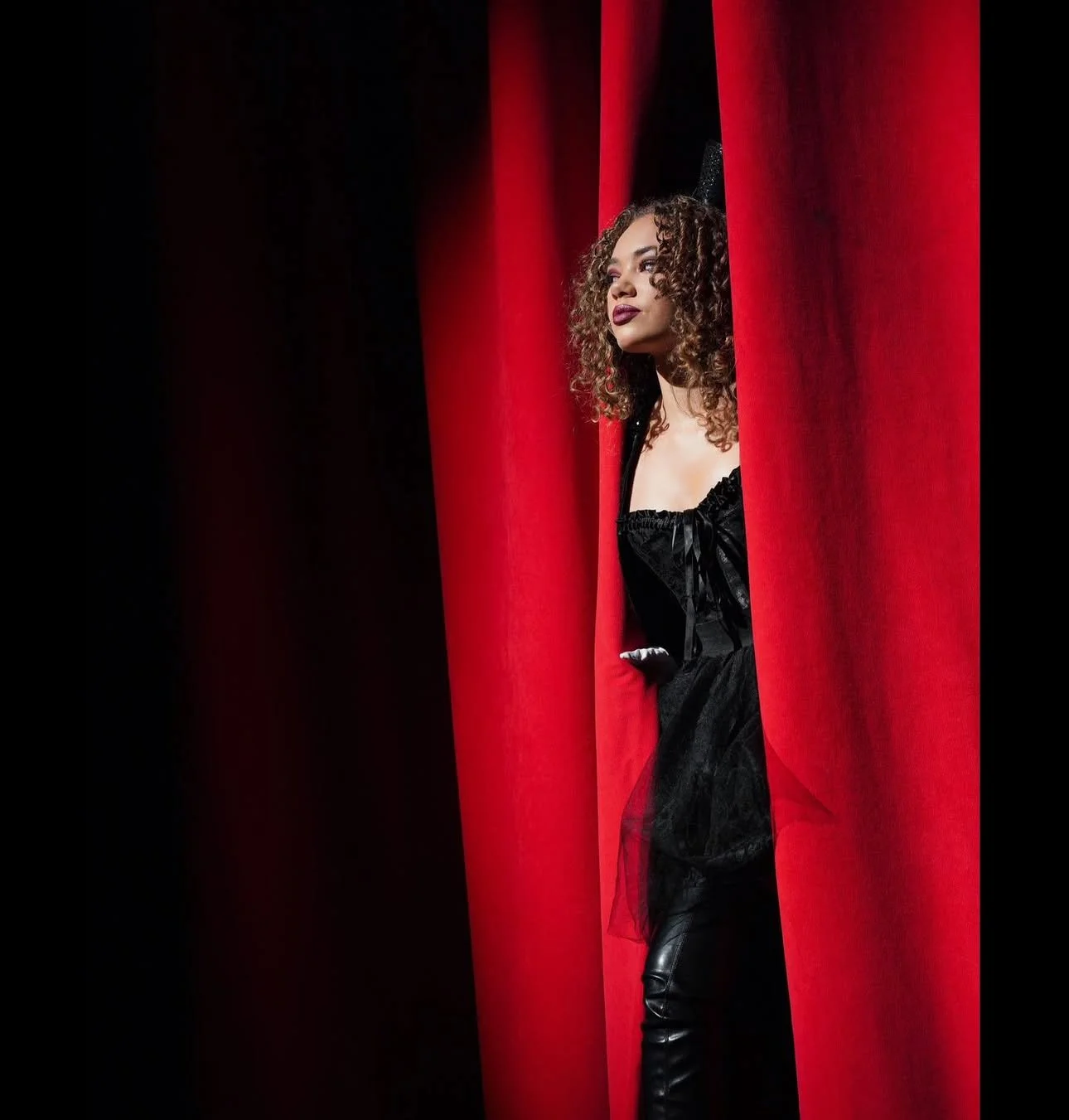 A woman with curly hair peeking through red theater curtains, wearing a black dress and looking thoughtfully into the distance.