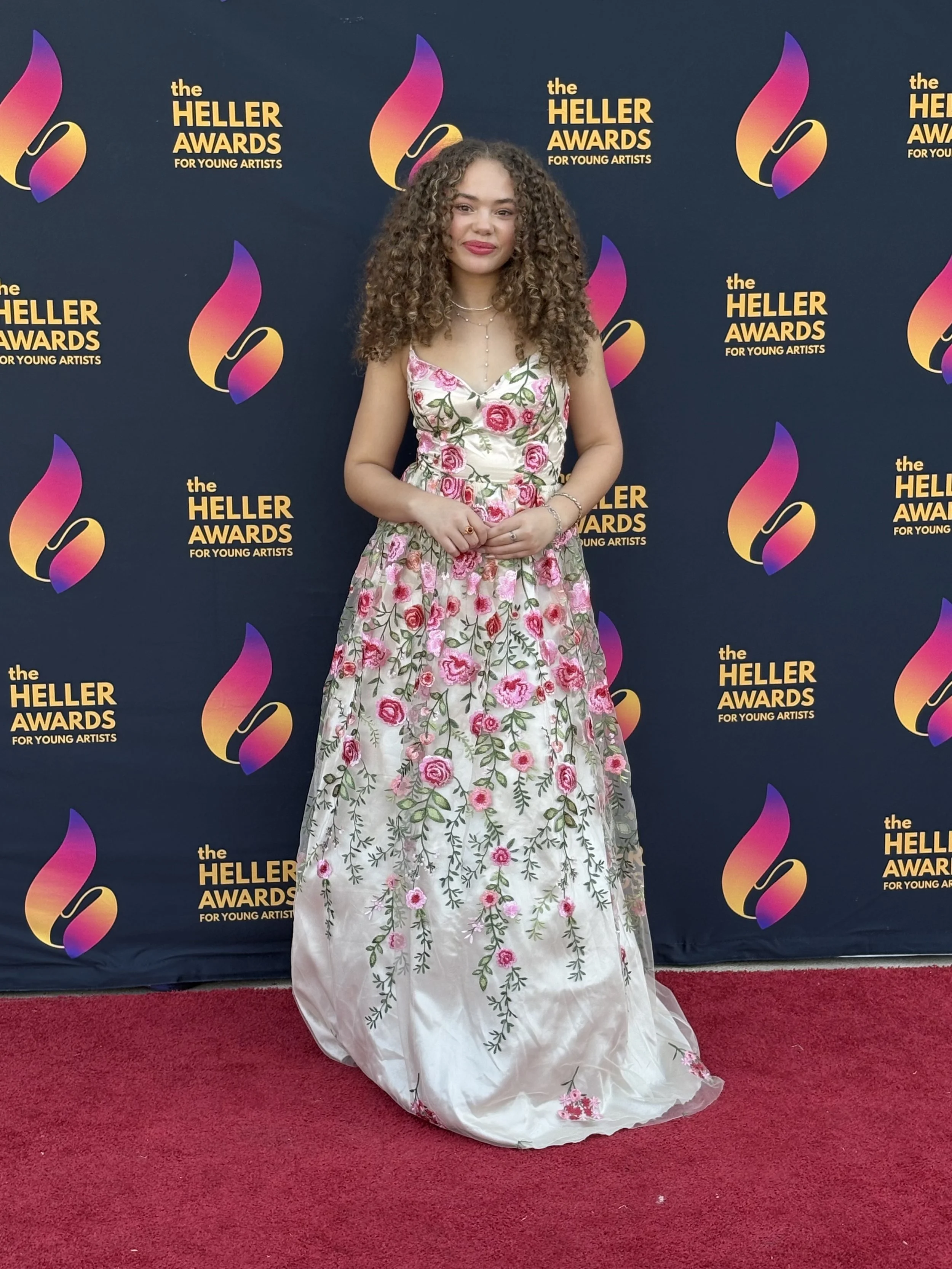Young woman with curly hair in a floral dress standing on a red carpet at the Heller Awards with a dark backdrop featuring the event's logo.