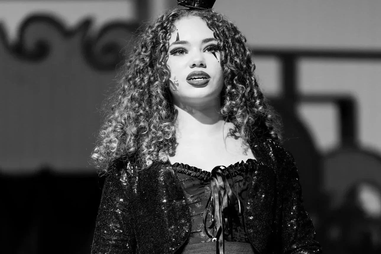 A woman with curly hair and makeup, wearing a sparkly black top with a ribbon on the front, standing on stage.