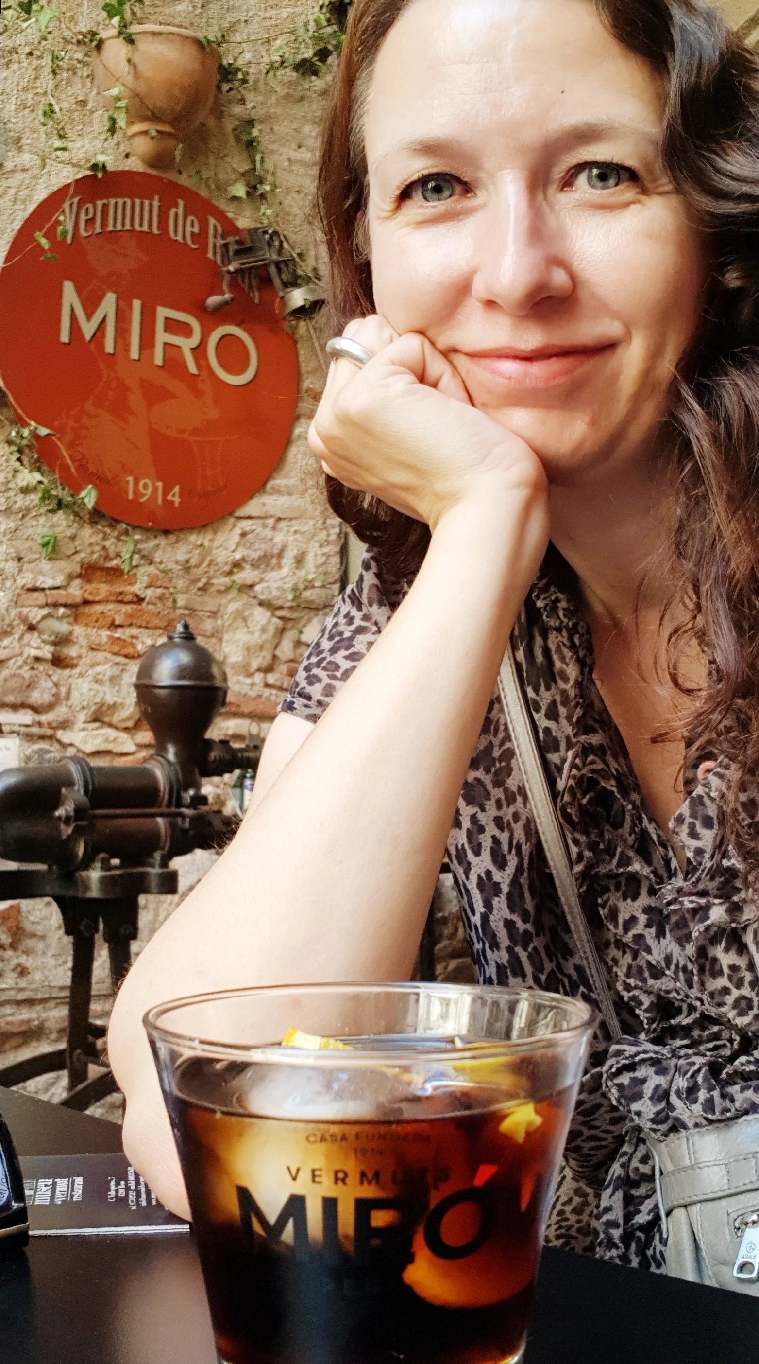 Kvindelig person sidder ved bord med glas med cola og citrusskiver, baggrund med murstensvæg og et skilt med teksten 'Vermut de Rive Miro 1914'.