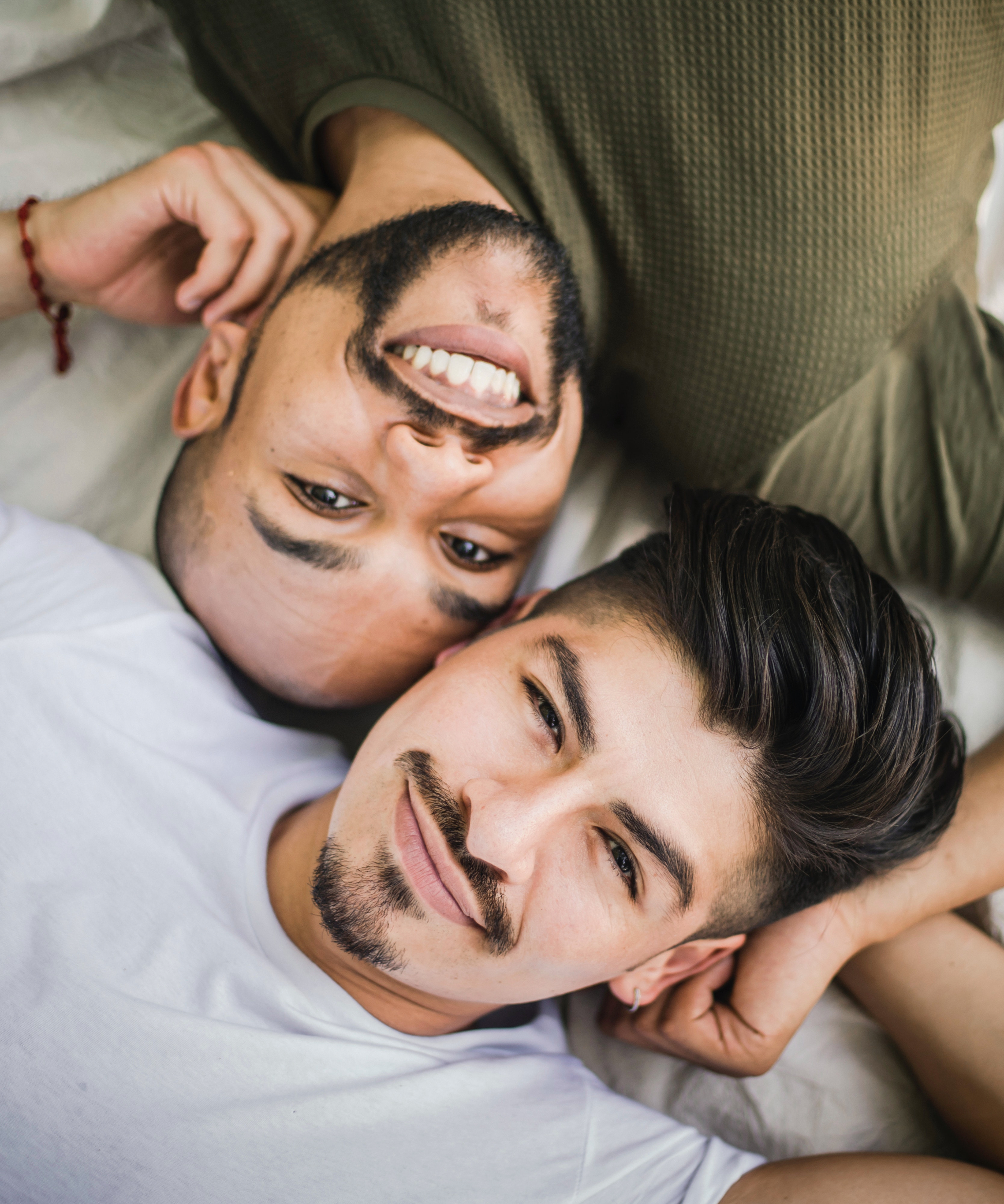 Two men lying on a bed, smiling, with their heads close together