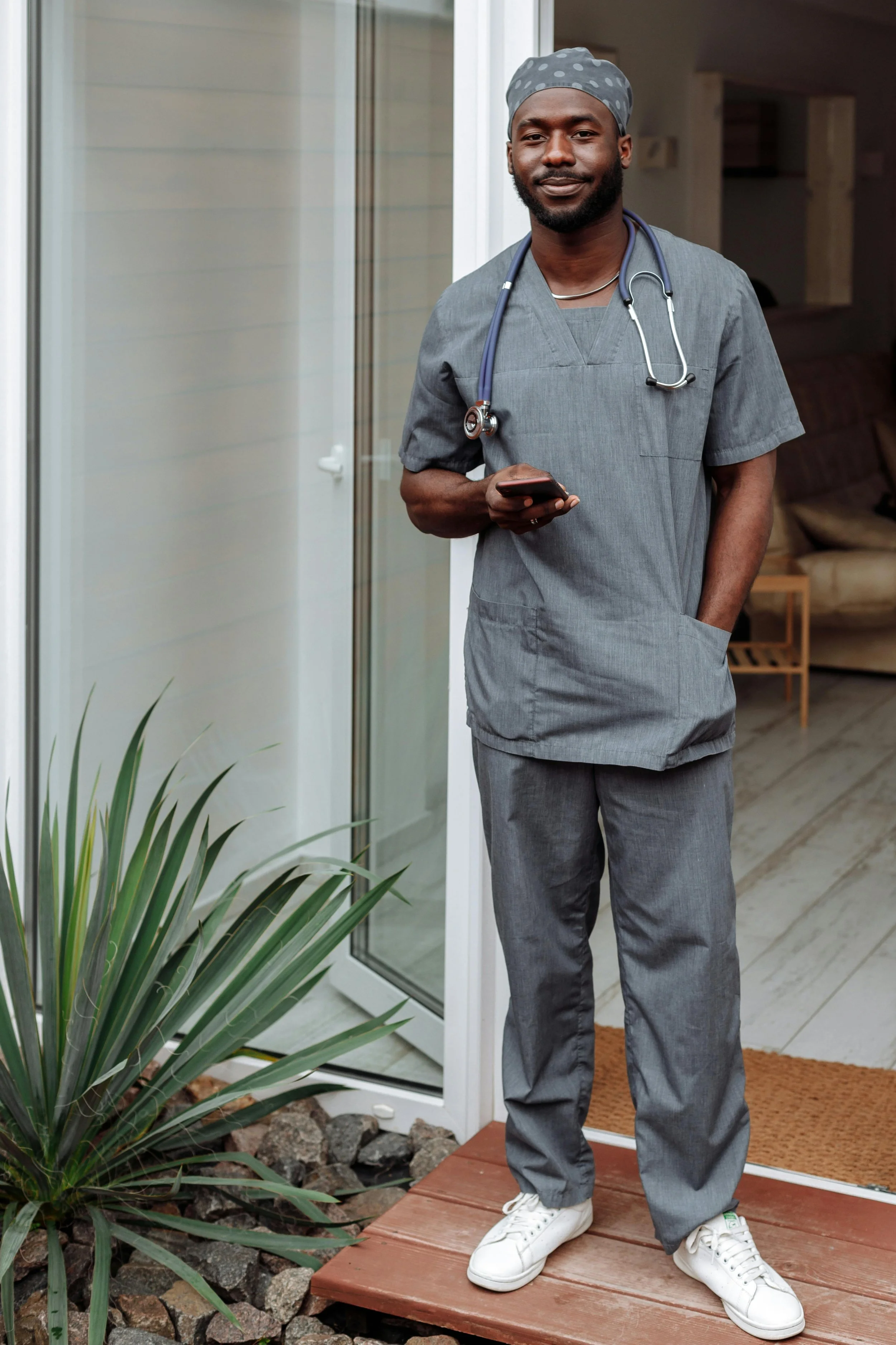 A male healthcare worker, dressed in gray scrubs with a stethoscope around his neck, standing on a porch and looking at his phone