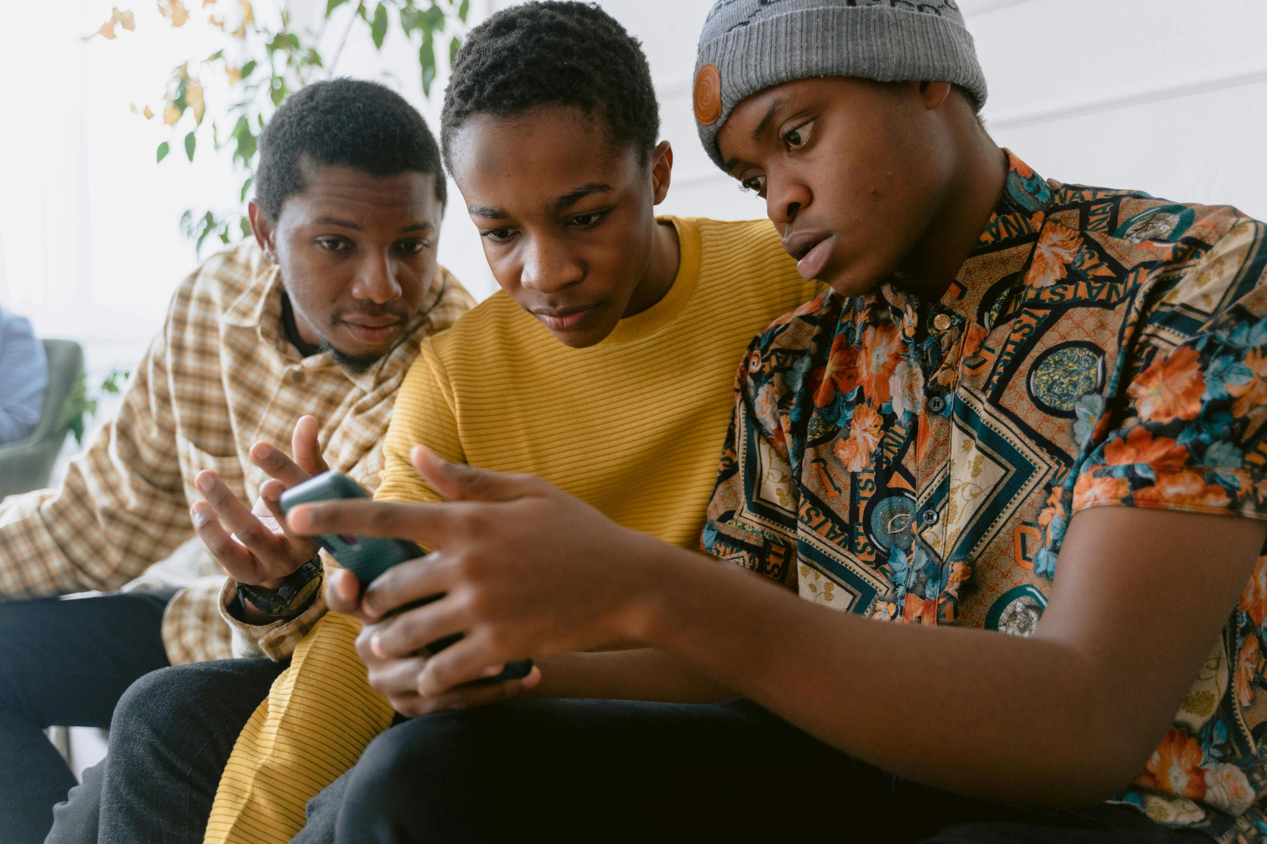 Three young men sit closely together looking at a smartphone held by one of them, indoors with a window and some greenery in the background.