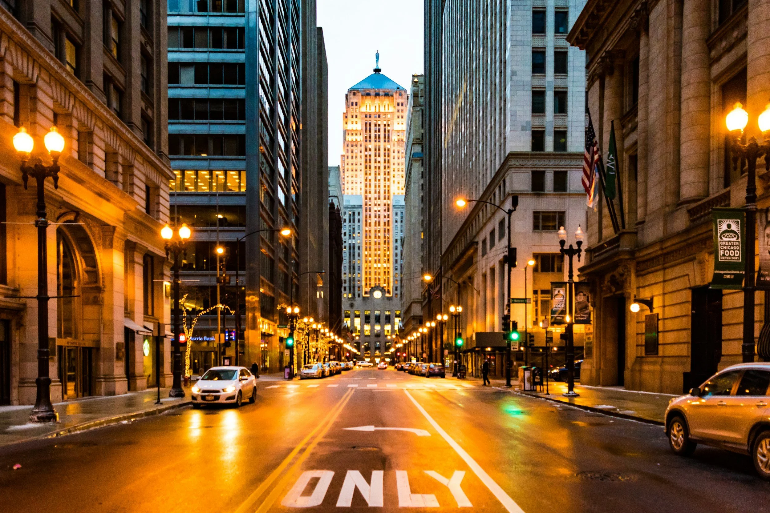 Lasalle Street in Chicago at dusk with tall buildings, lit street lamps, traffic, and an American flag, with the Illinois Tower in the background.
