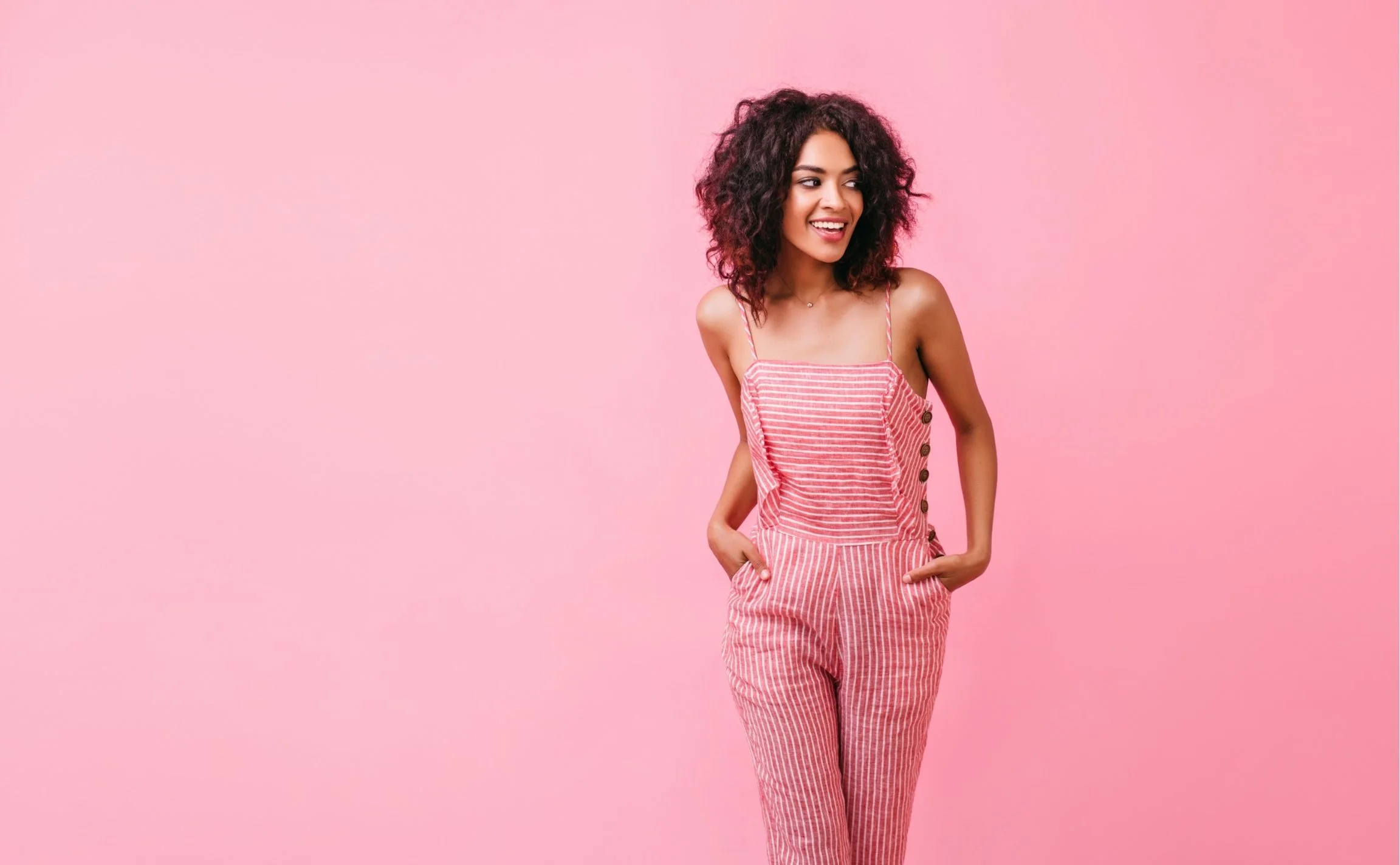Young woman smiling, wearing pink and white striped jumpsuit with buttons on the side, standing against pink background.