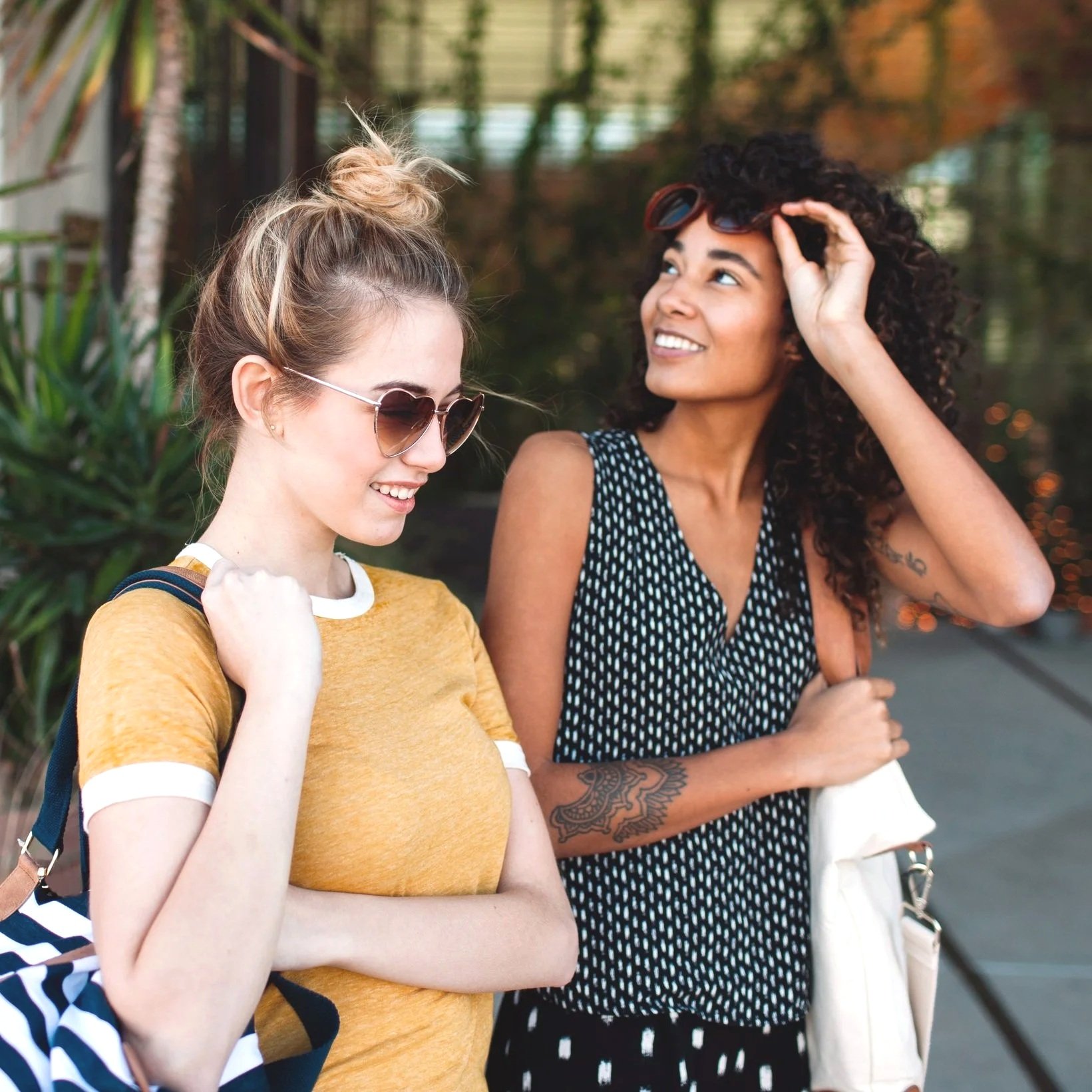 Two women, one with blonde hair and sunglasses wearing a yellow shirt, and the other with curly black hair wearing a black and white polka dot dress, standing outdoors and smiling at each other.