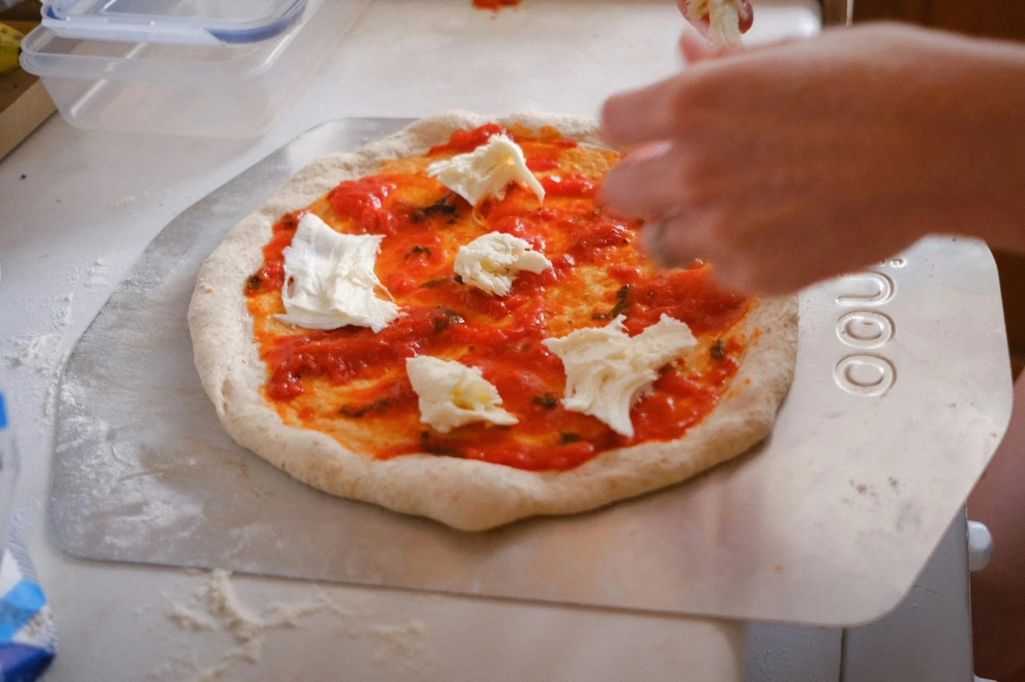 Eine Person fügt Ricotta-Käse auf eine gerade frisch belegte Pizza mit Tomatensauce und frischen Kräutern.