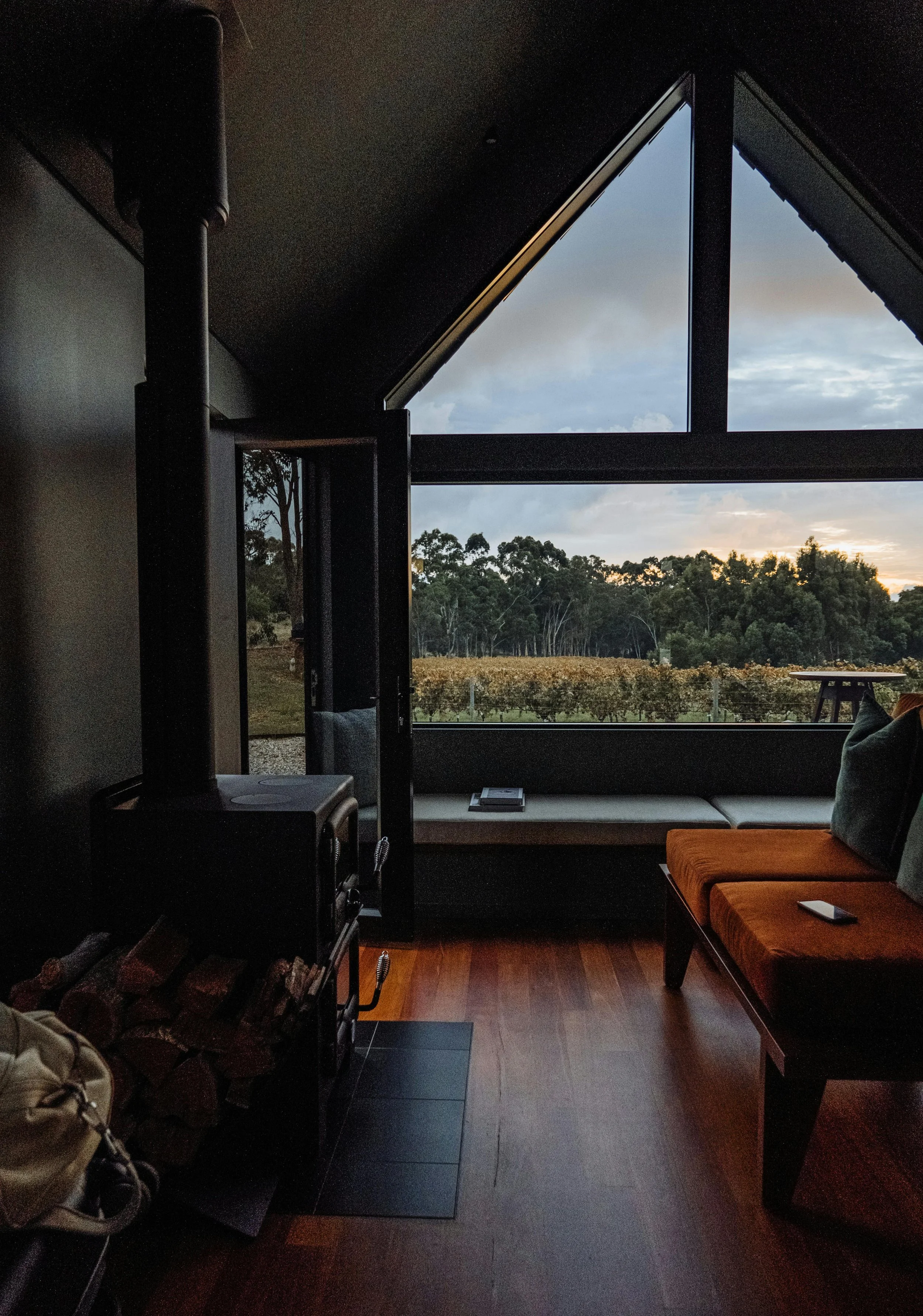 Interior of a cozy living room with a large triangular window showing an outdoor landscape of trees and a vineyard at sunset.